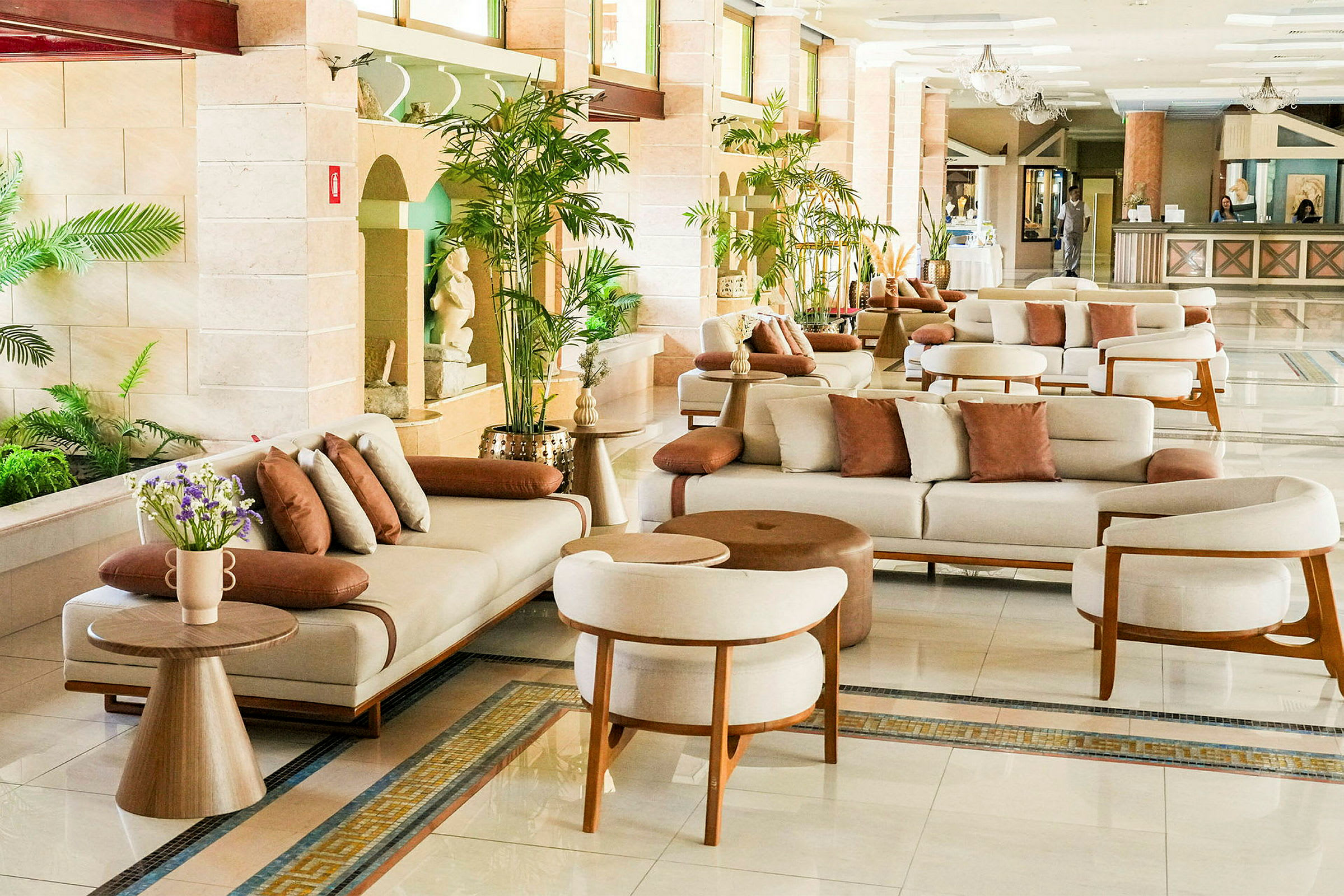 Et hotellobbyområde med hvide sofaer, brune puder og små træborde, omgivet af frodige planter. (AI-genereret tekst)