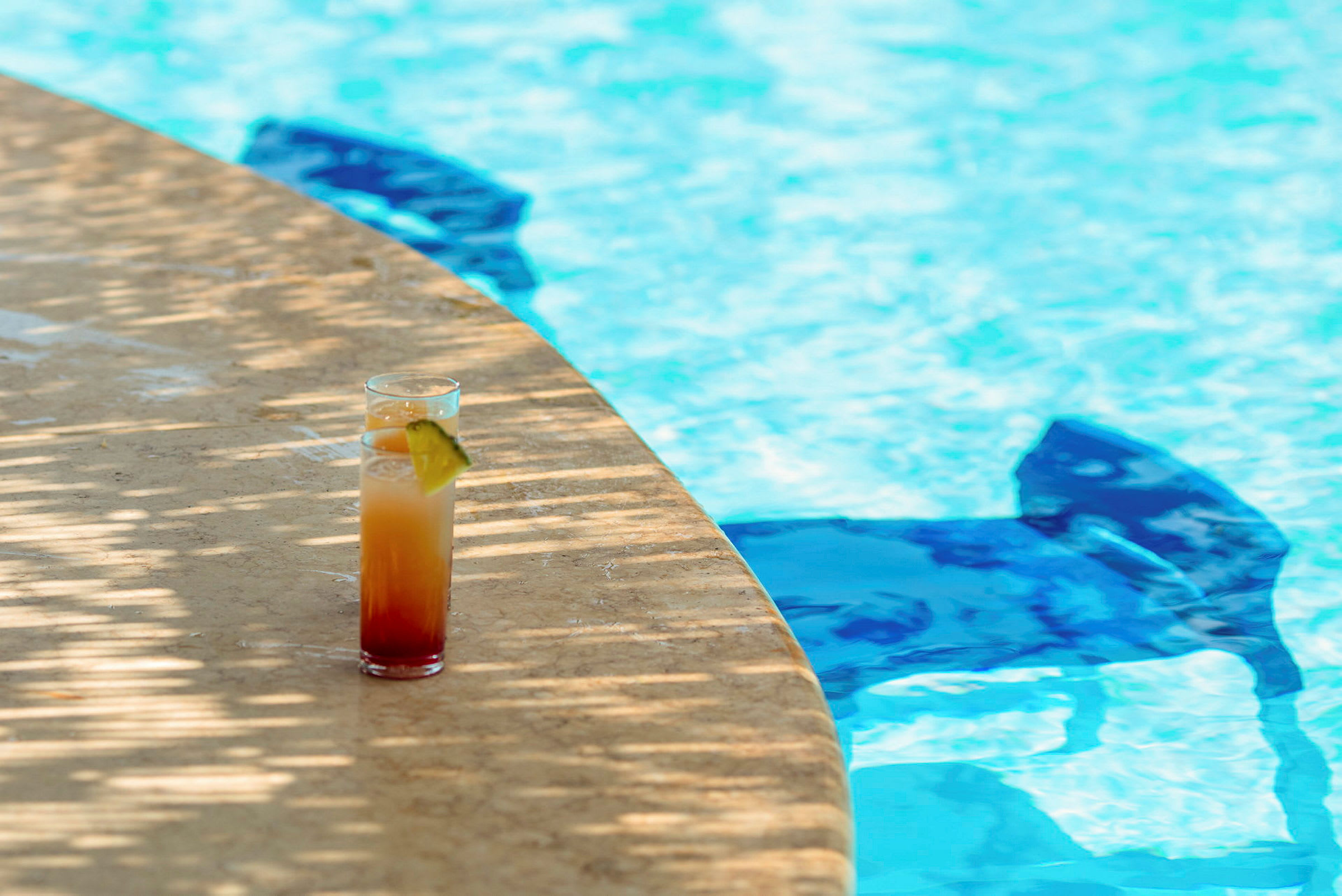 Et glas med en farverig drink står på en stenkant ved siden af en swimmingpool. (AI-genereret tekst)