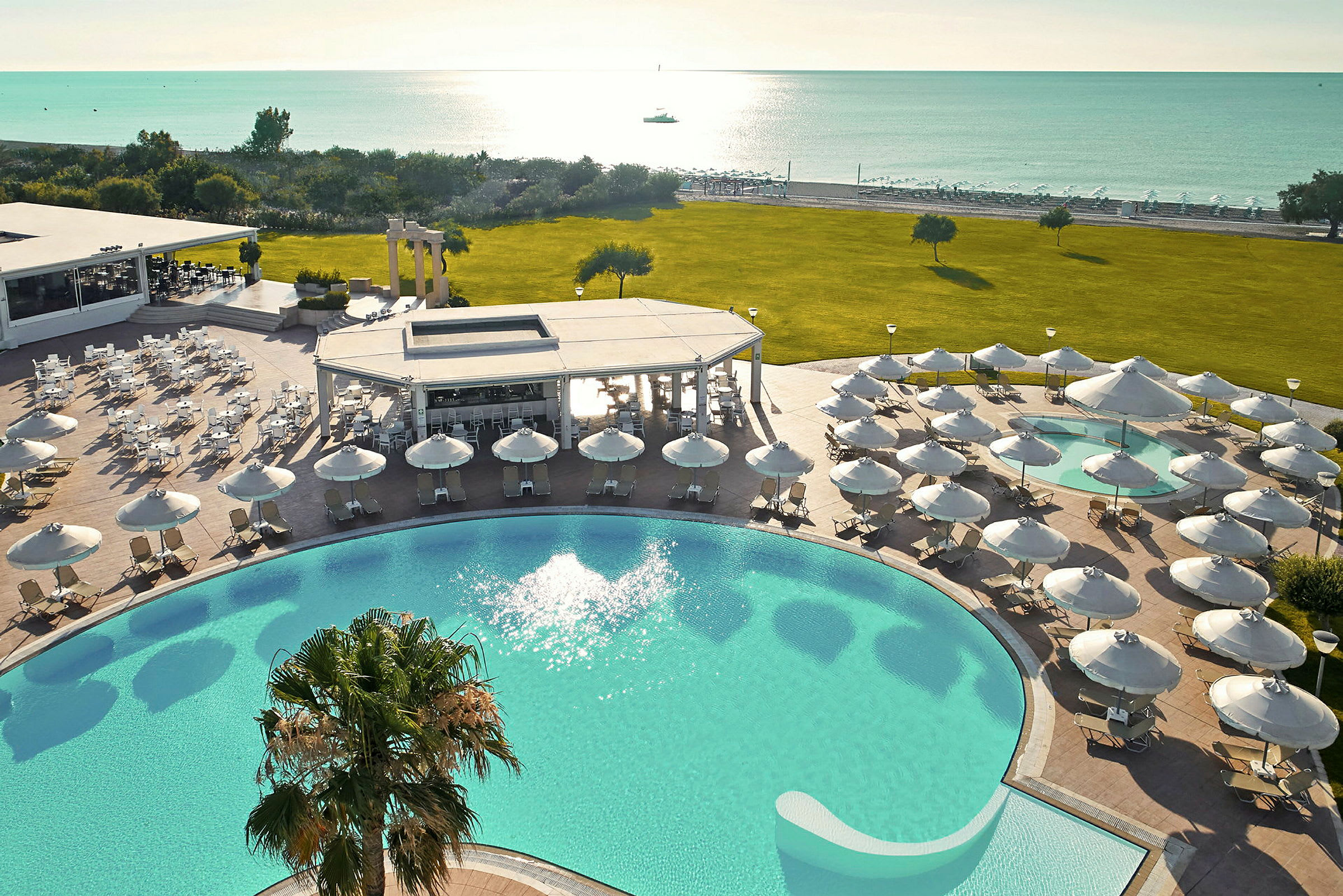 En udsigt over et solrigt resort med pool, parasoller, grøn græsplæne og havudsigt. (AI-genereret tekst)