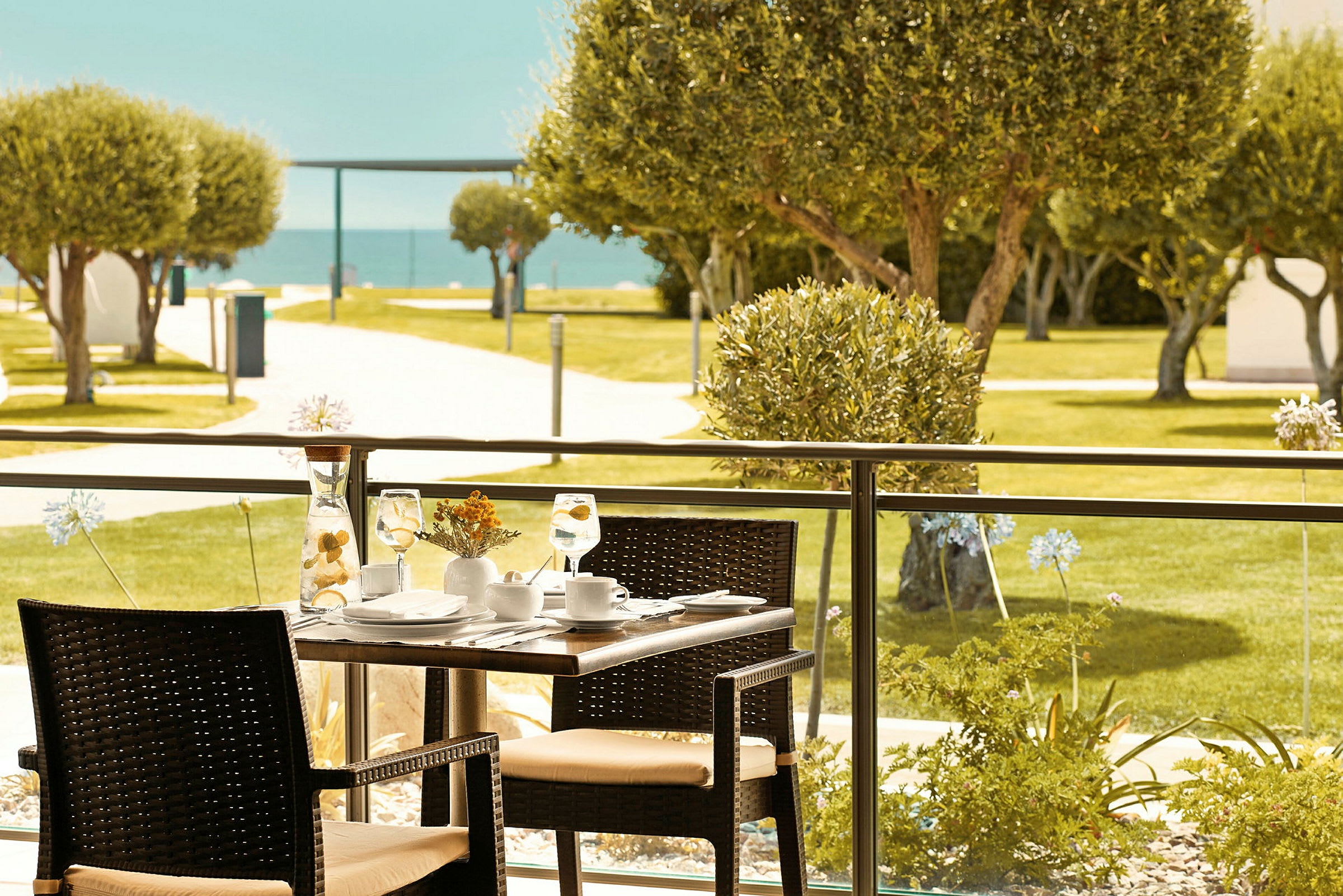 Et dækket bord med stole på en udendørs terrasse med udsigt over grønne områder og havet. (AI-genereret tekst)