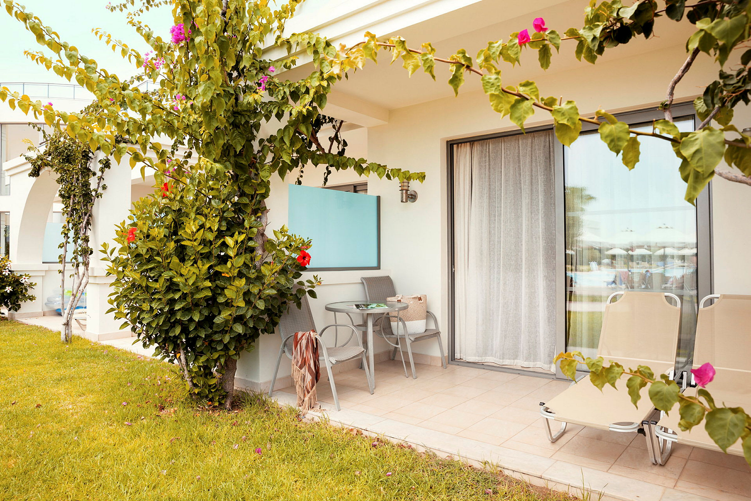 Terrasse med bord, stole og liggestole. Foran er græsplæne og blomstrende planter. (AI-genereret tekst)