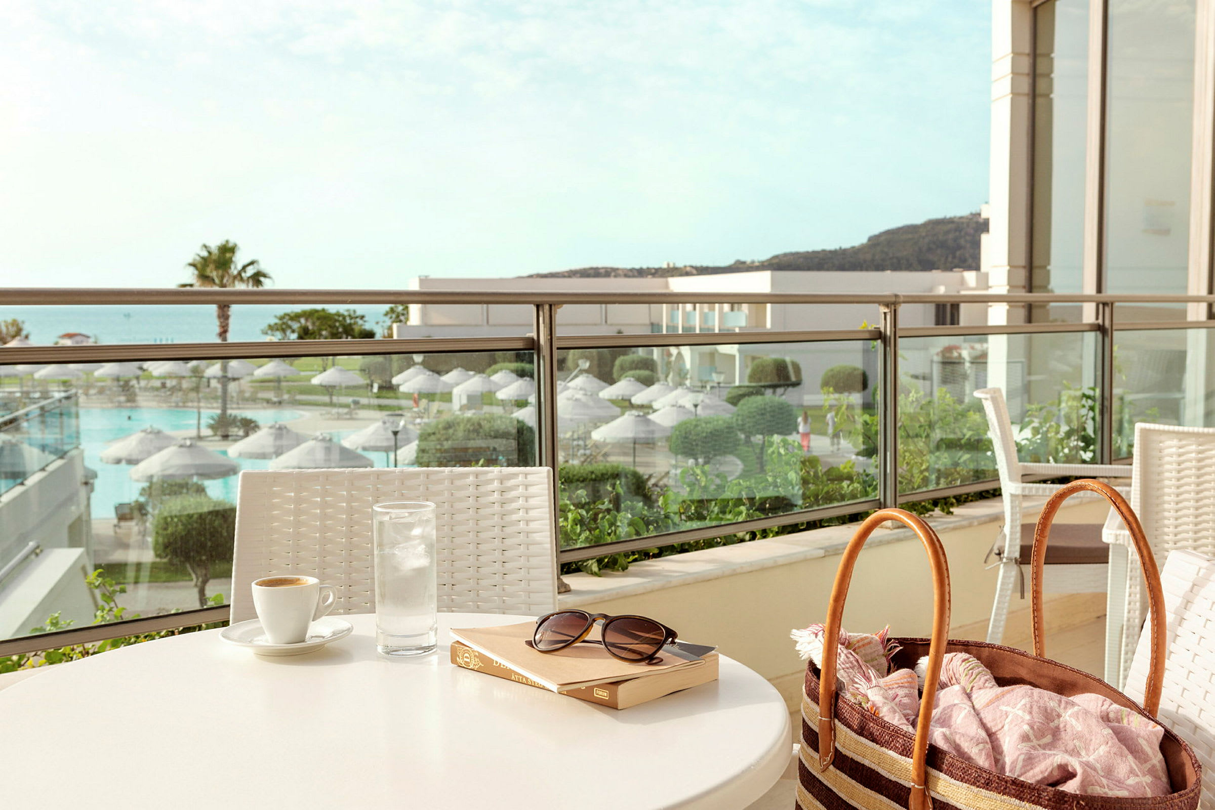 Et bord med en kop kaffe, et glas vand og en bog placeret på en terrasse med udsigt over et poolområde og beplantning. (AI-genereret tekst)