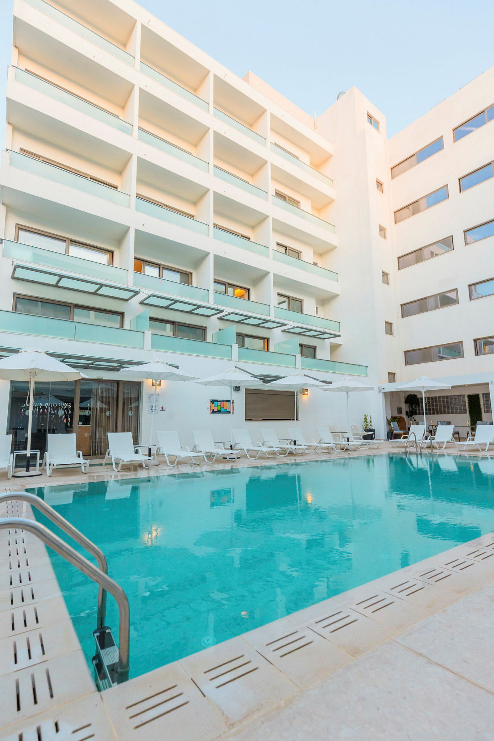 En hotelpool omgivet af liggestole og en moderne hotelbygning. (AI-genereret tekst)