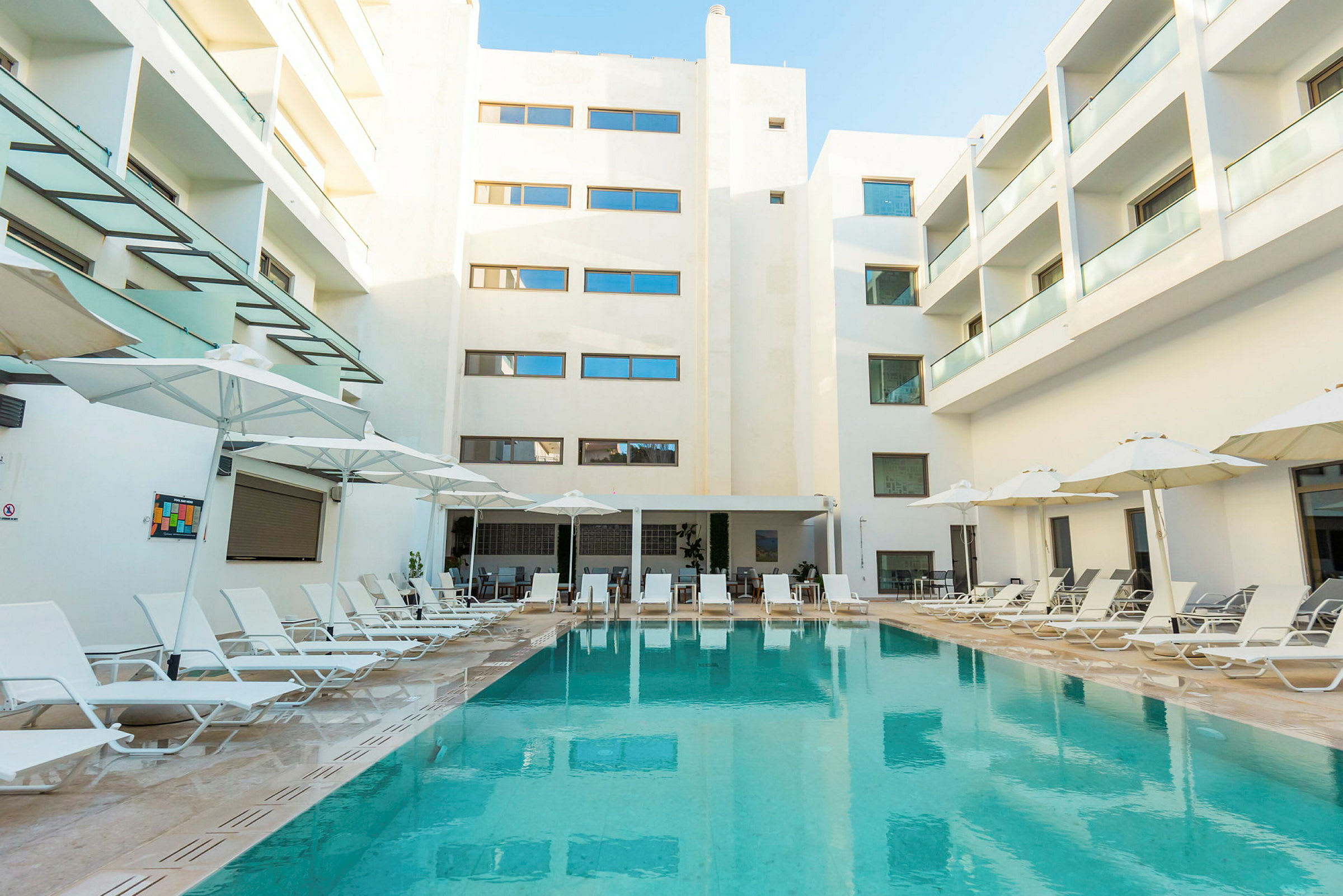 Billede af et hotelområde med en swimmingpool omgivet af liggestole og parasoller ovenover. Baggrunden viser hvide bygninger med altaner. (AI-genereret tekst)