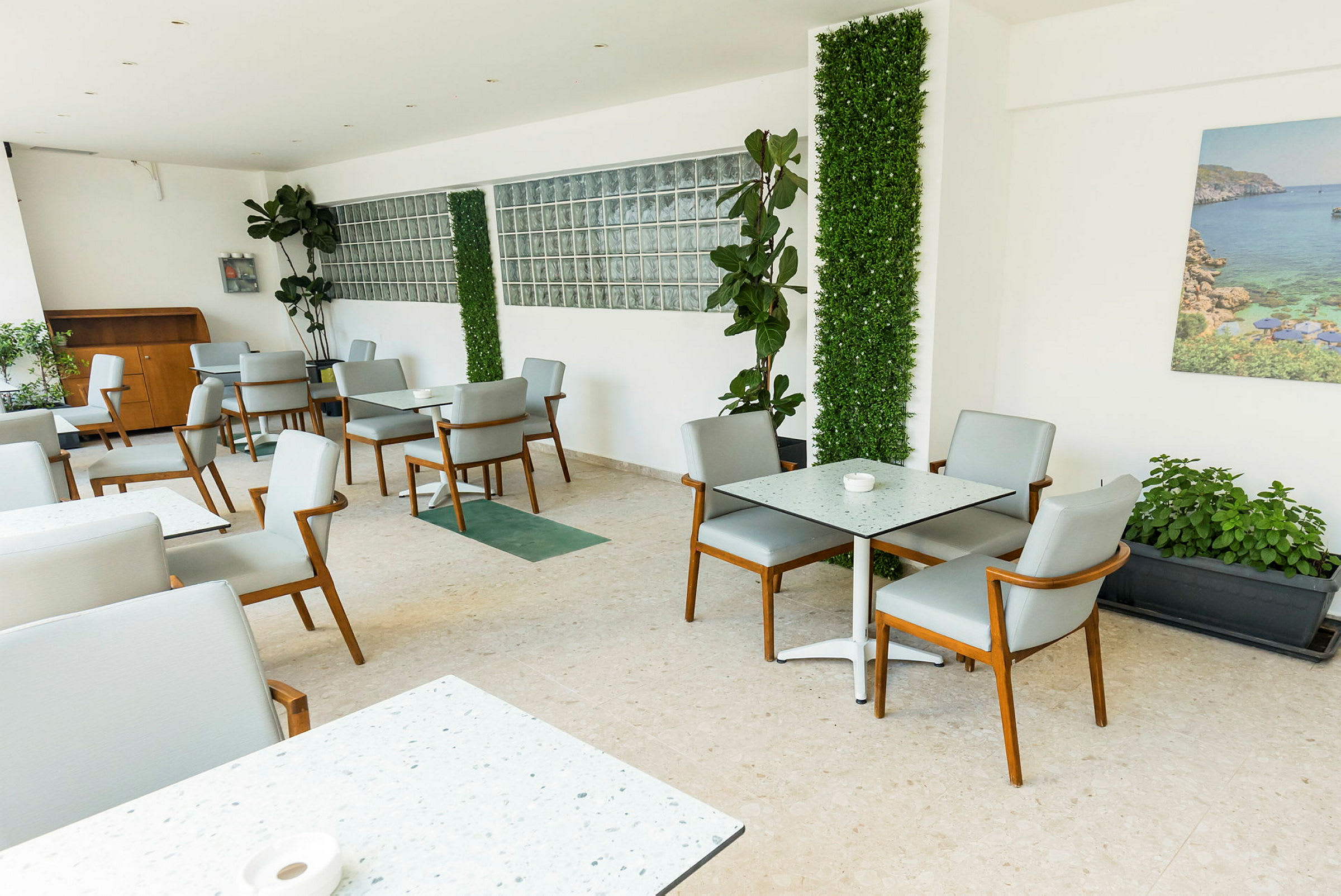Et moderne caféområde med borde og stole, grønne planter på væggene og et fotografi af et kystlandskab, der hænger på væggen. (AI-genereret tekst)