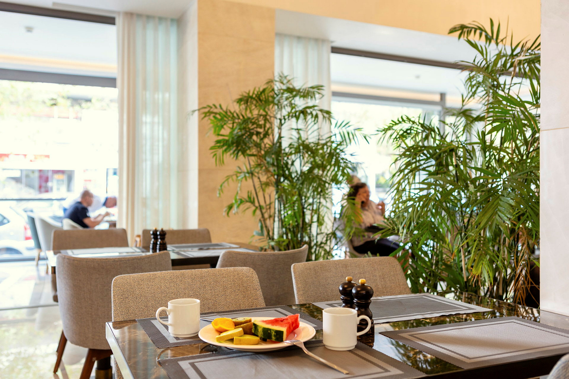 En café med borde dækket med en tallerken indeholdende frugt, kopper og krydderbeholdere, omgivet af grøn vegetation. (AI-genereret tekst)