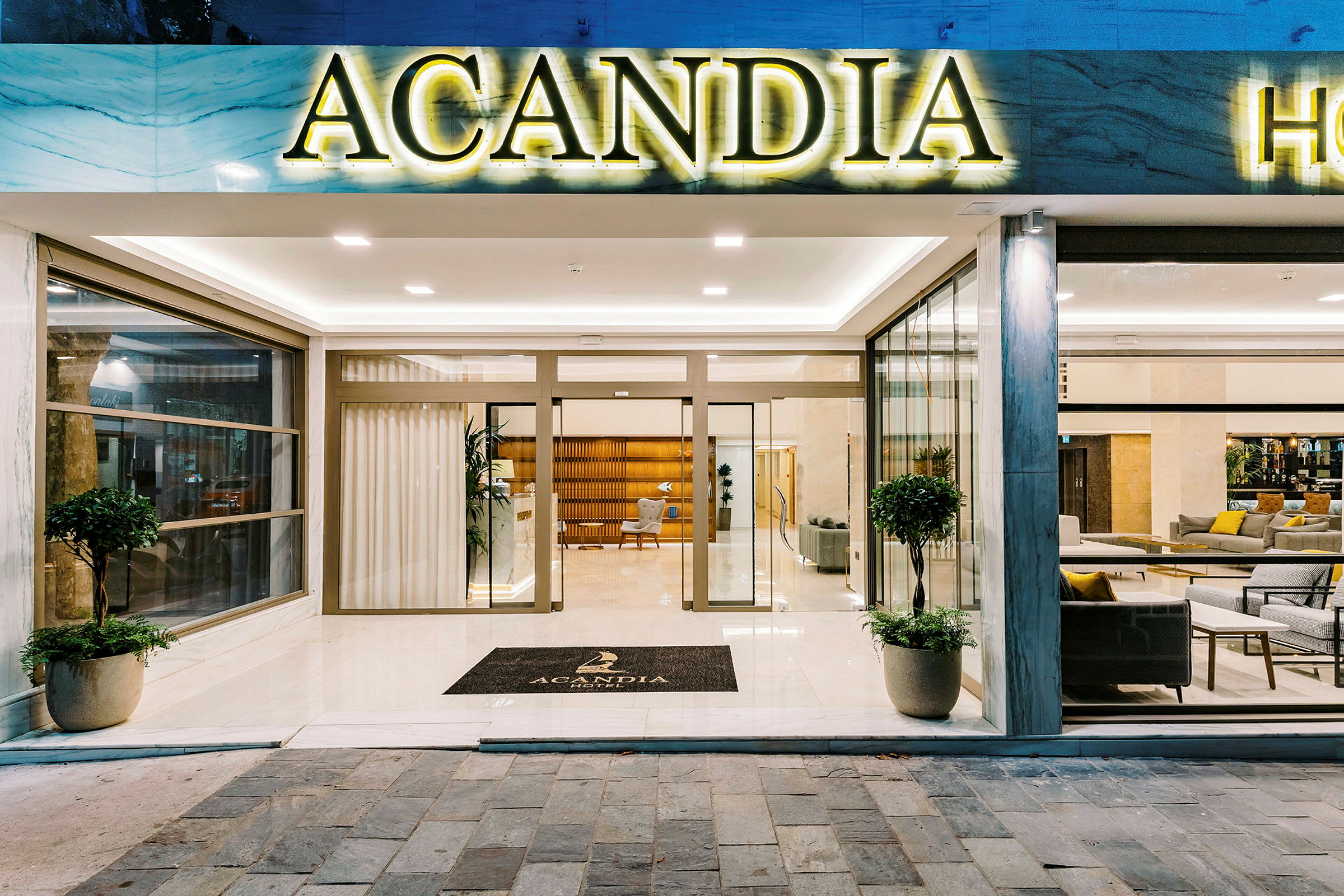 Indgangen til Hotel Acandia med glasdøre og et skilt med hotellets navn. (AI-genereret tekst)