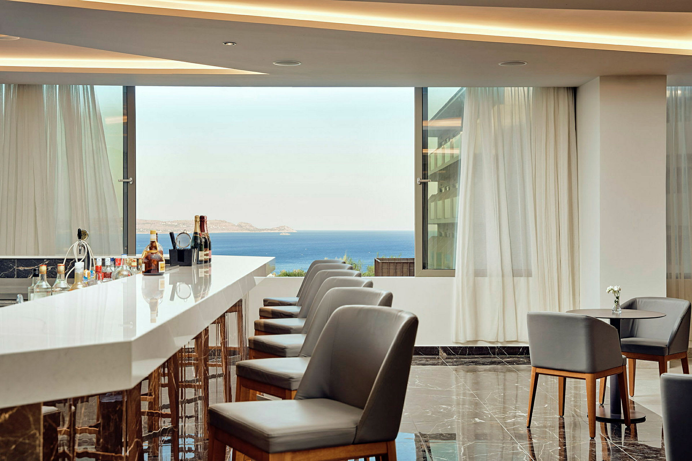 En hotelbar med moderne stole, marmorgulve og udsigt over havet fra store vinduer. (AI-genereret tekst)