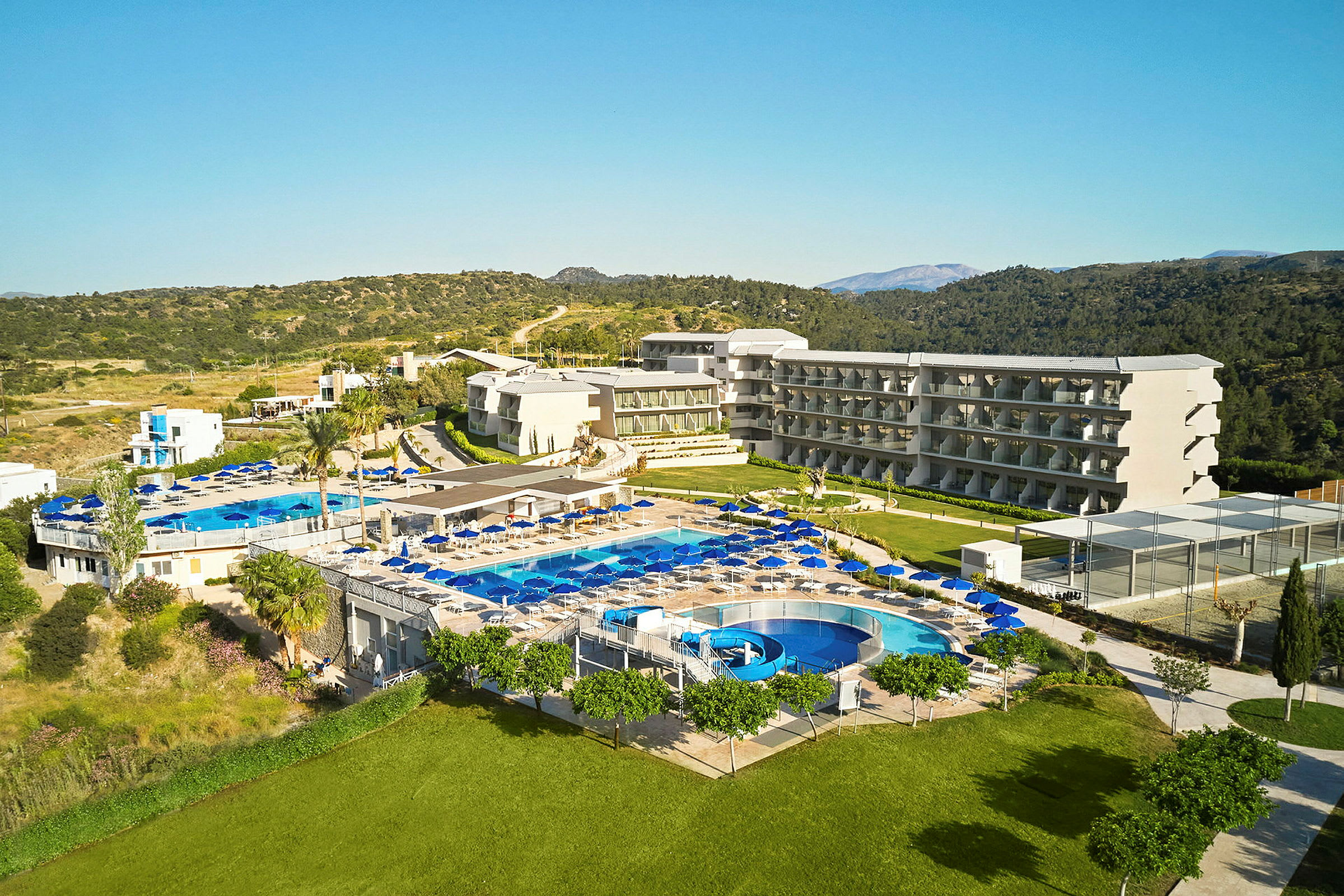 En udsigt over et hotelområde med flere bygninger, swimmingpools og solvogne, omgivet af grønt. (AI-genereret tekst)