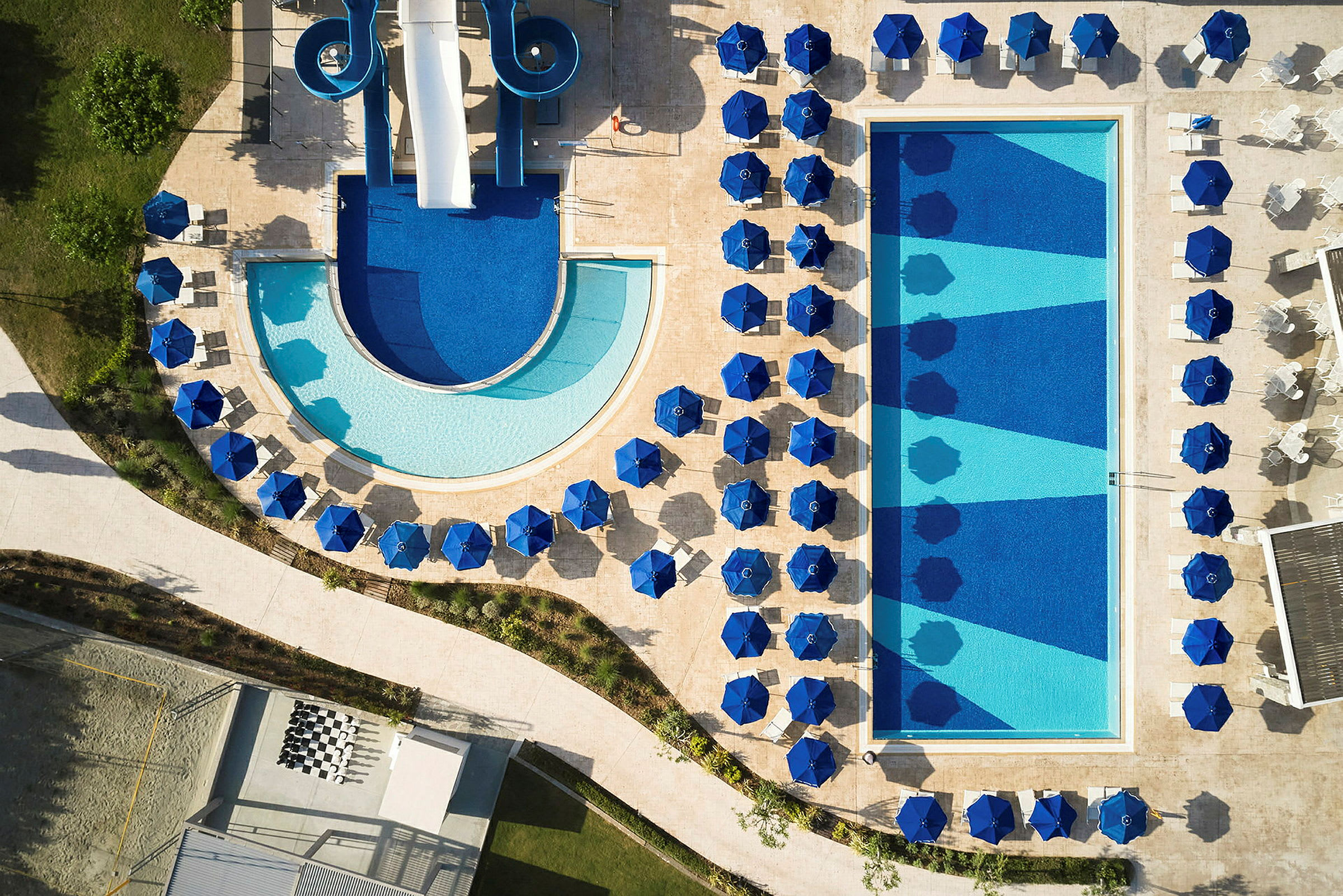 Luftfoto af en swimmingpool med blå parasoller og liggestole omkring. (AI-genereret tekst)