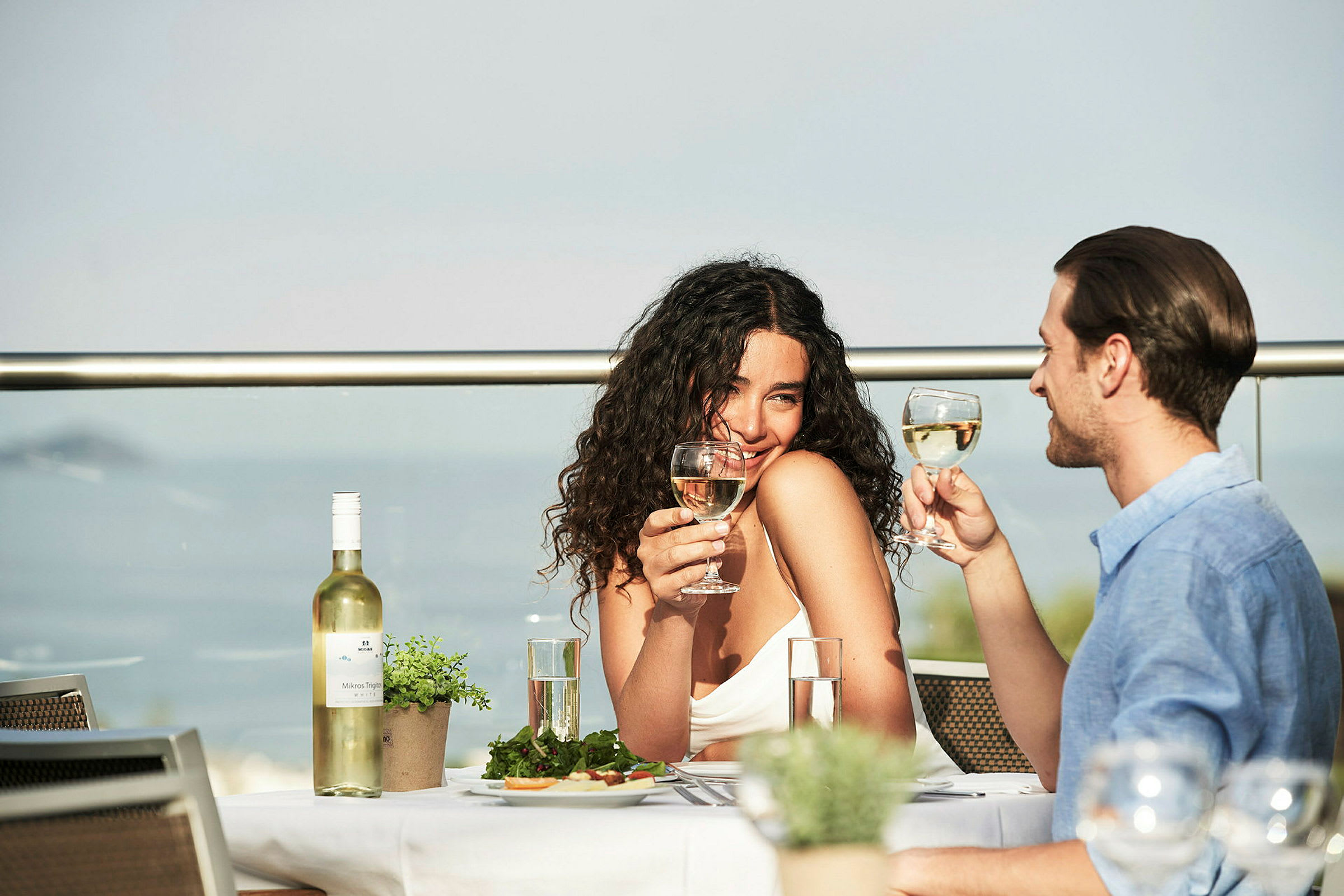 Personer skåler med vinglas ved et dækket bord på en terrasse. (AI-genereret tekst)
