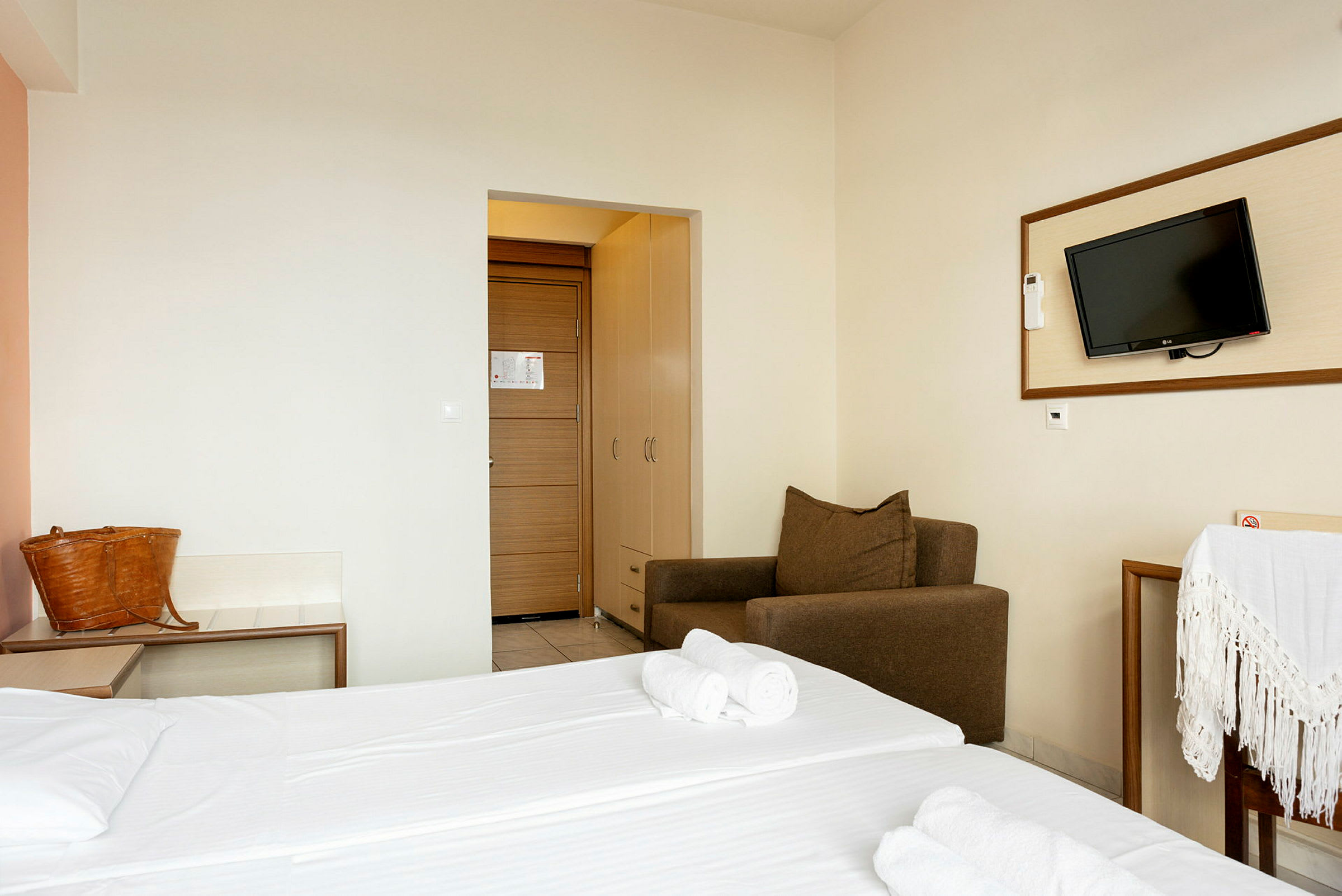 Hotelværelse med dobbeltseng, brun lænestol, væghængt TV og dør med garderobe. (AI-genereret tekst)