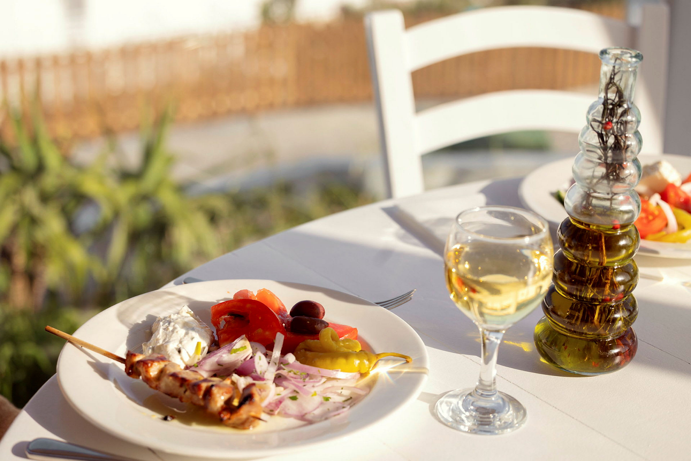 En tallerken med grillspyd, grøntsager og sauce på et bord udendørs, med et glas hvidvin og en flaske olivenolie. (AI-genereret tekst)