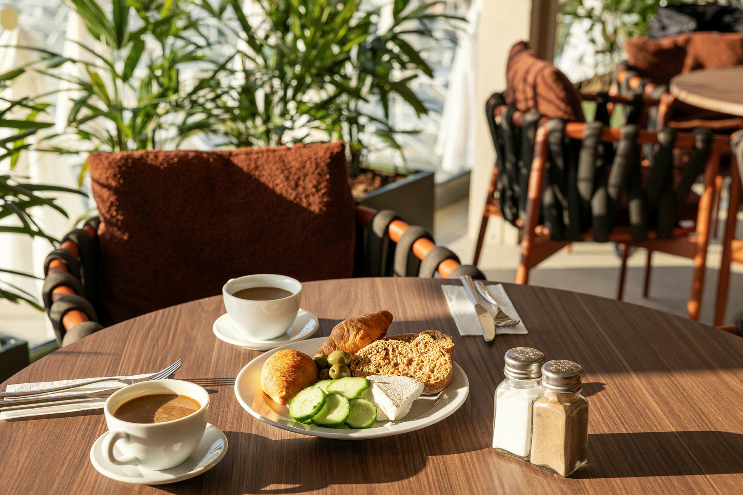 En morgenmadssætning med kaffe og en tallerken med brød, ost, agurkeskiver og en croissant på et bord. (AI-genereret tekst)