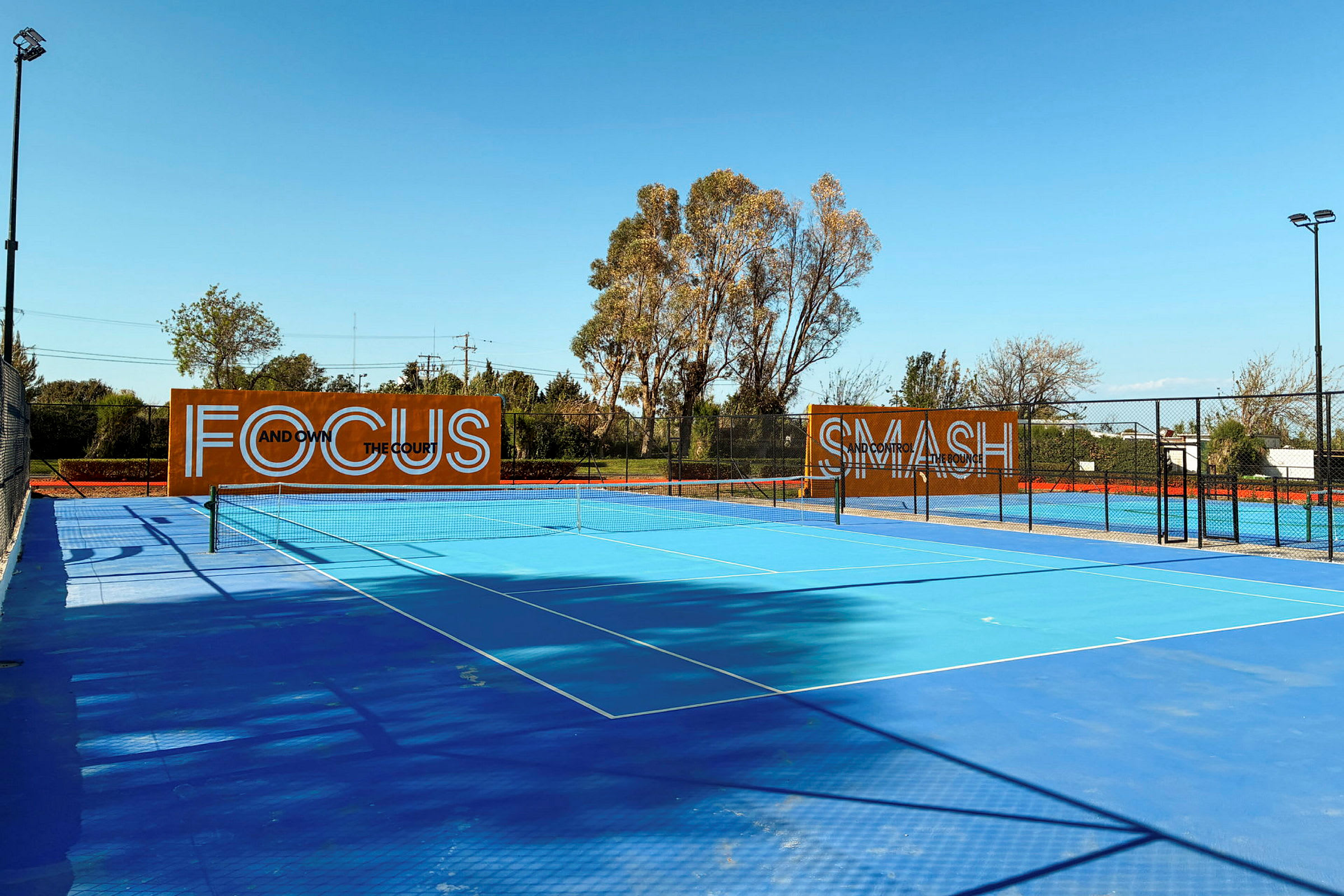 Blå tennisbane udendørs med træer i baggrunden og skilte med teksten "Focus" og "Smash". (AI-genereret tekst)