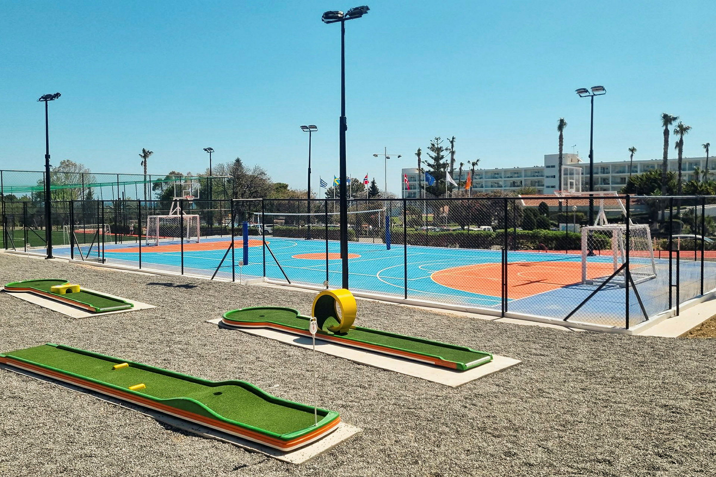 Et billede af et udendørsmiljø med en minigolfbane i forgrunden og en farverig sportsbane med orange og blå overflade omgivet af et hegn i baggrunden. (AI-genereret tekst)