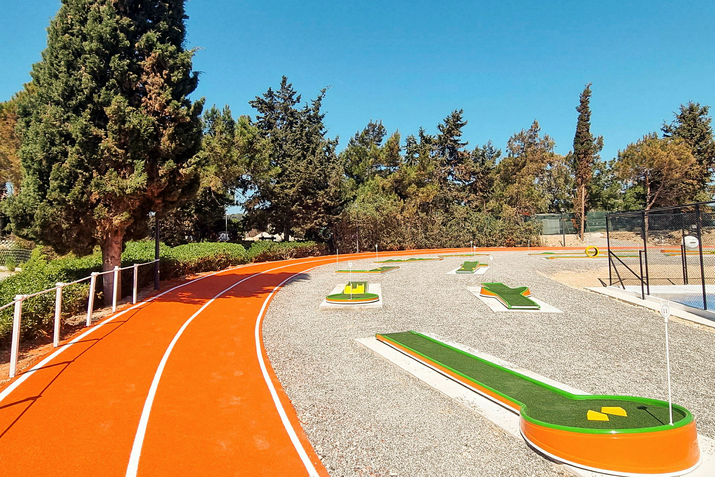 Et billede af en minigolfbane ved siden af en orange løbebane. Billedet er taget udendørs med grøn vegetation og klar blå himmel i baggrunden. (AI-genereret tekst)