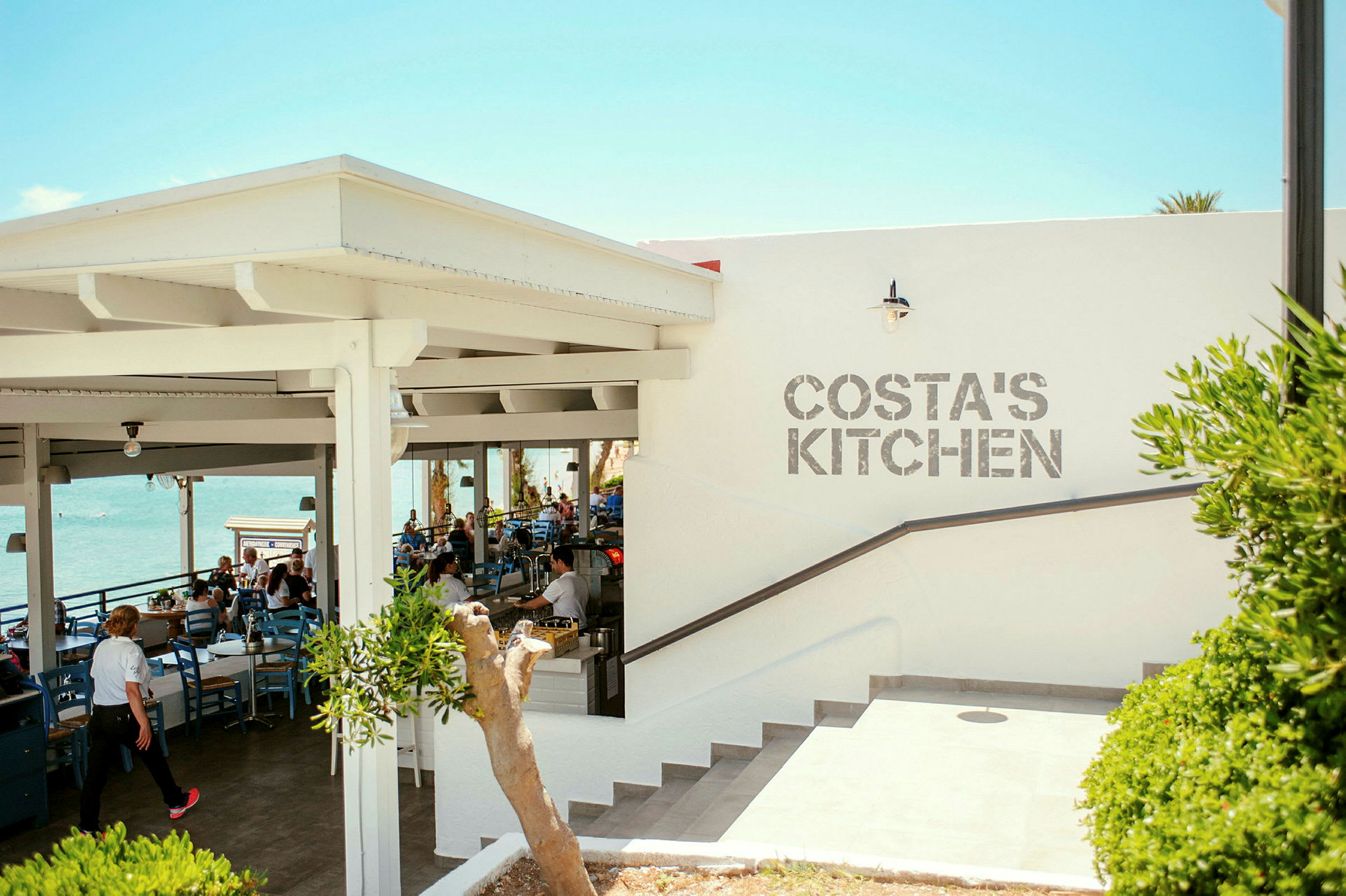 En udendørs restaurant med havudsigt og teksten 'Costa's Kitchen' på væggen. (AI-genereret tekst)