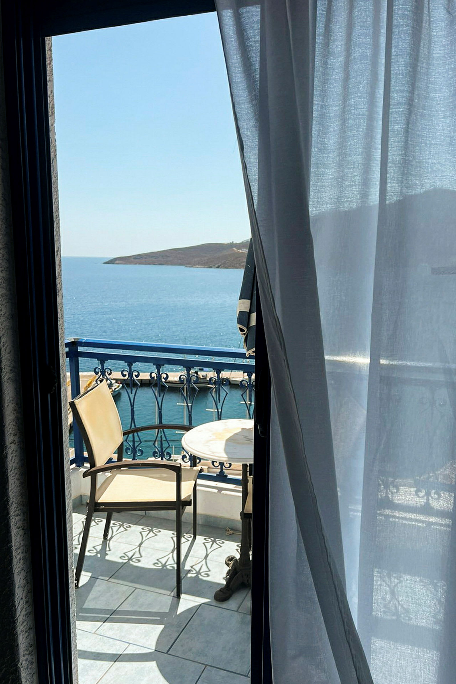 Udsigt fra et værelse mod en balkon med to stole og et bord samt hav og kyst i baggrunden. (AI-genereret tekst)