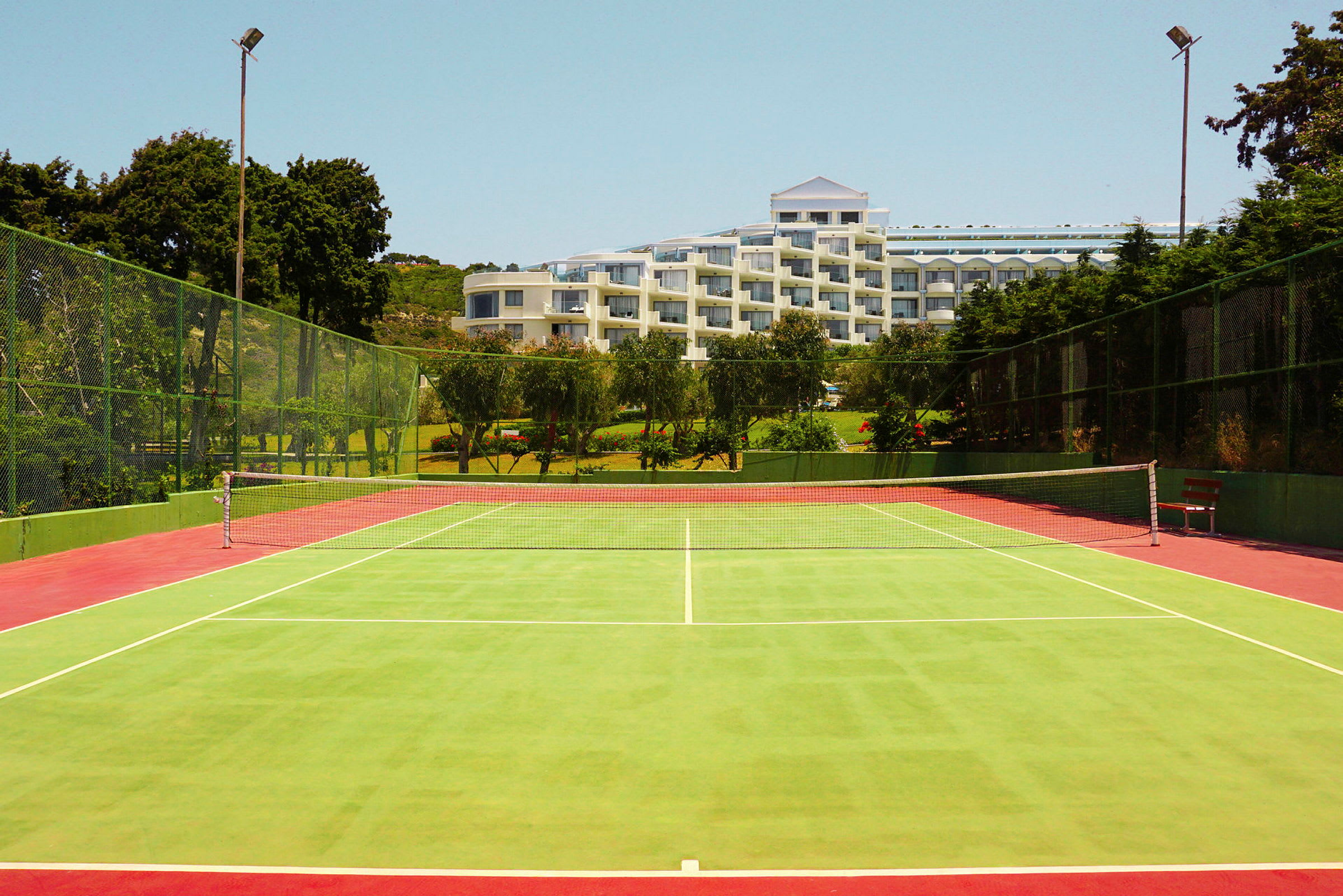 En grøn tennisbane med et net i midten og et hotel i baggrunden omgivet af træer. (AI-genereret tekst)