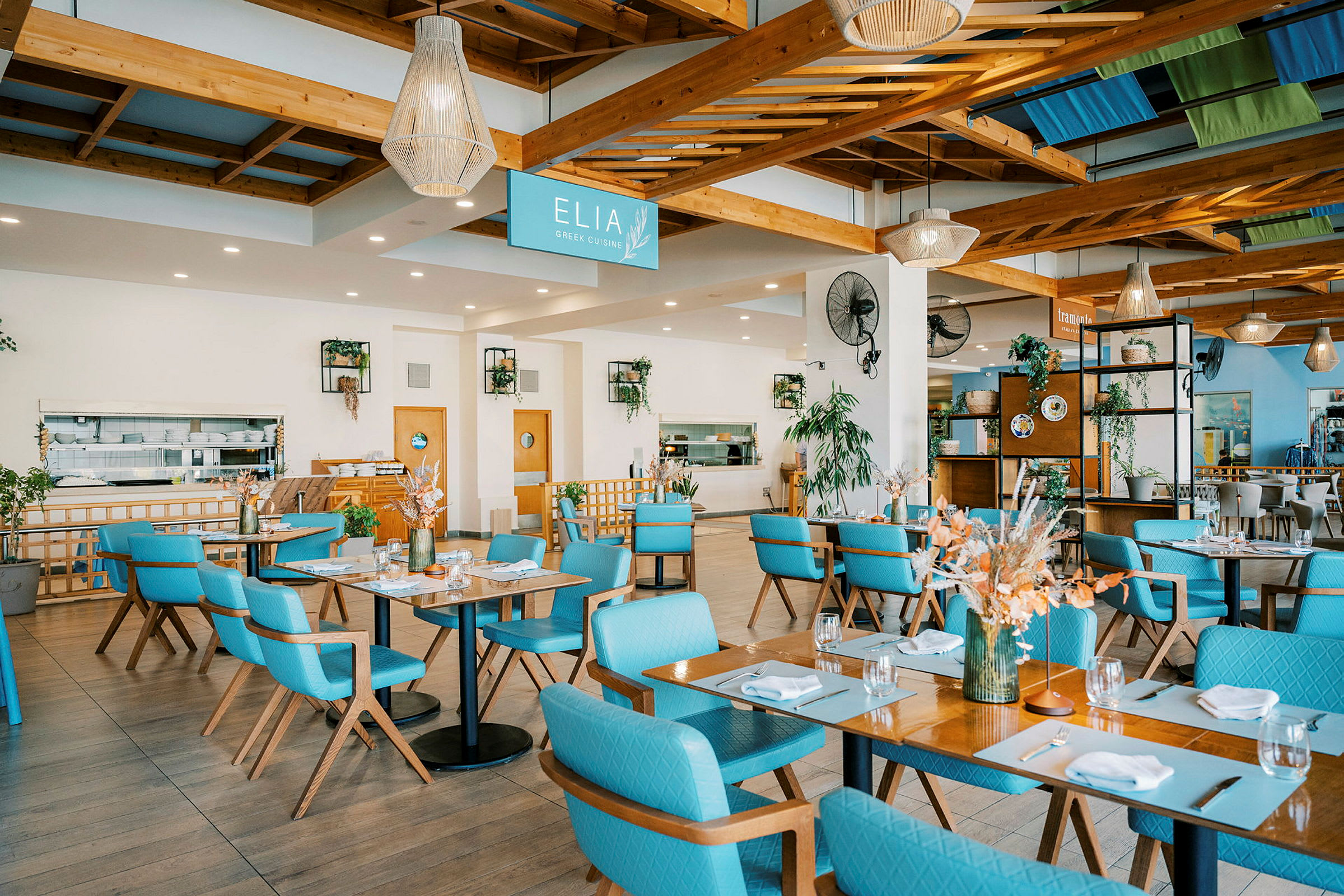 Et lyst og rummeligt restaurantmiljø med borde og blå stole. Dekorationer og grønne planter er en del af indretningen. (AI-genereret tekst)