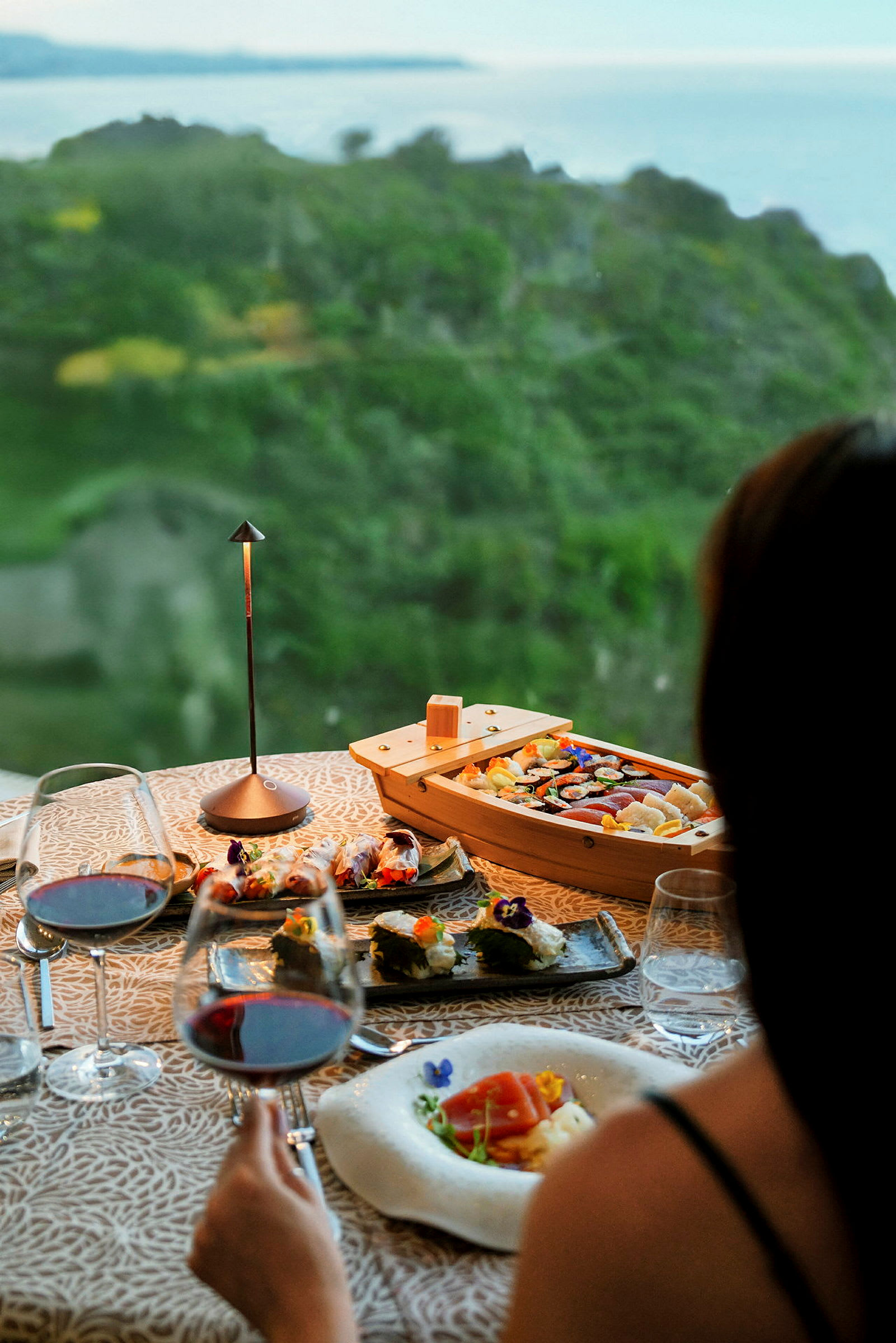 Et smukt opdækket måltid med sushi og vin står på et bord med udsigt over et grønt landskab og havet. (AI-genereret tekst)