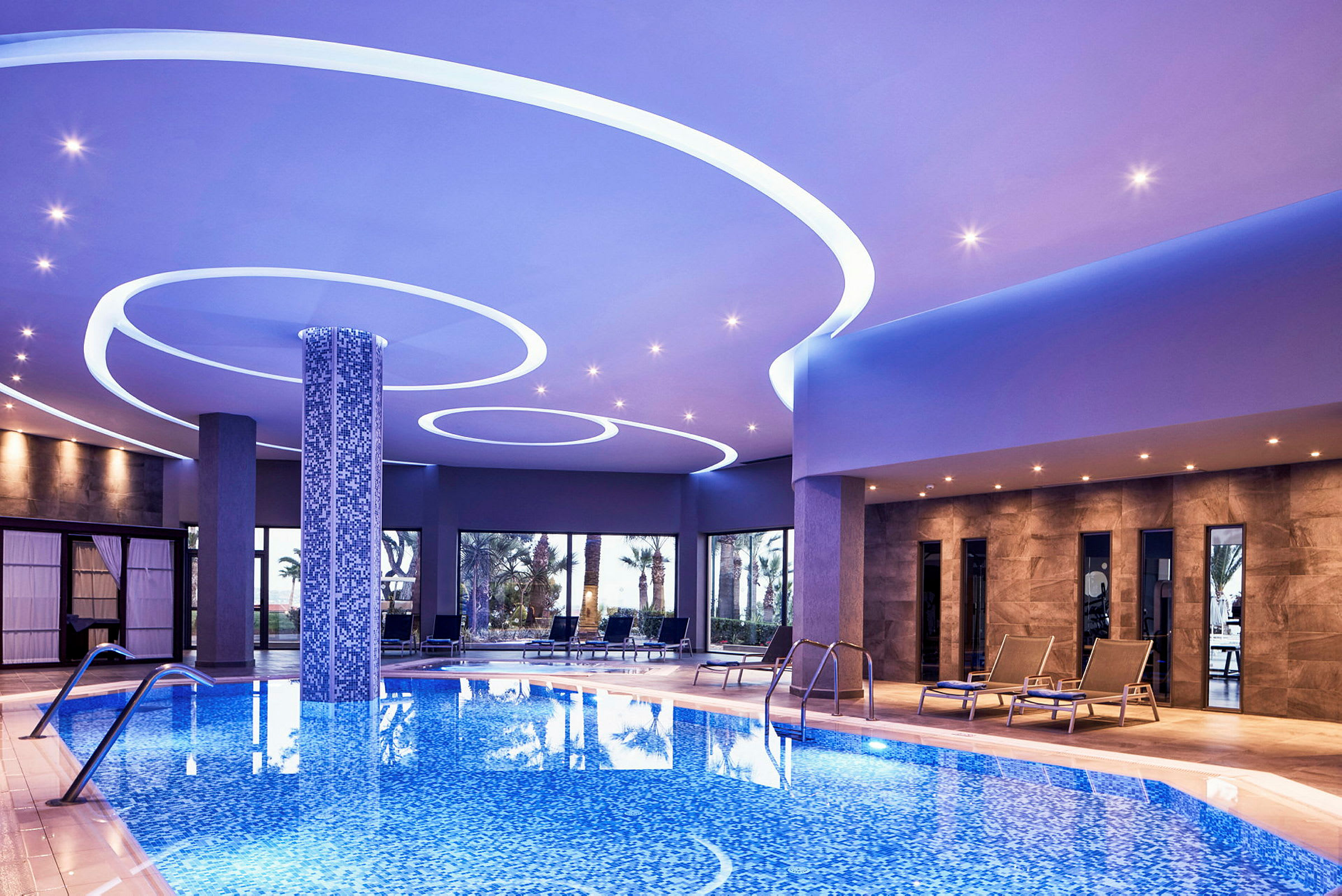 Indendørs pool med blå mosaikgulv og omkringliggende loungeområder. (AI-genereret tekst)