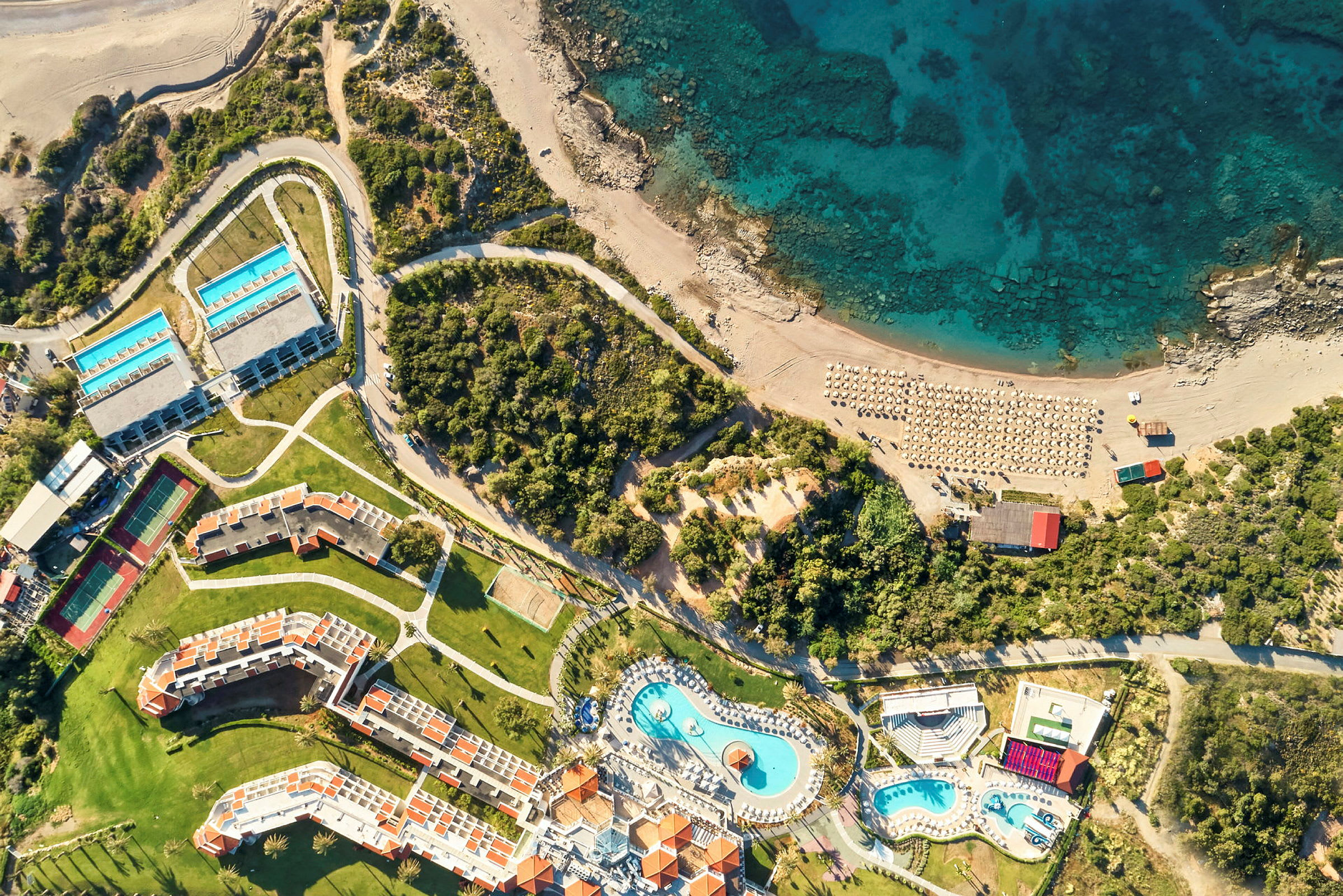 Luftfoto af et hotelområde med pools, bygninger, strand og hav. (AI-genereret tekst)