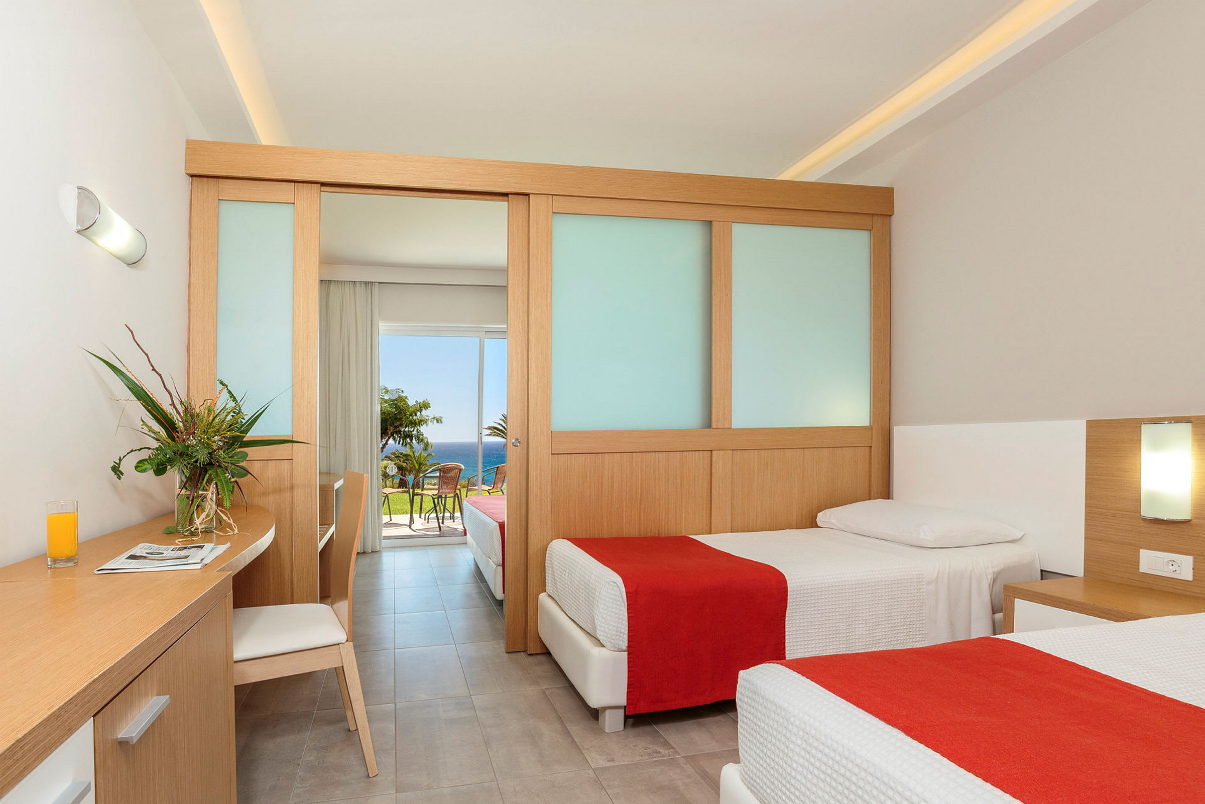 Et hotelværelse med to enkeltsenge, en arbejdsplads med en stol og en glasvase med blomster. Værelset har en skydedør, der fører til en balkon med udsigt over havet. (AI-genereret tekst)