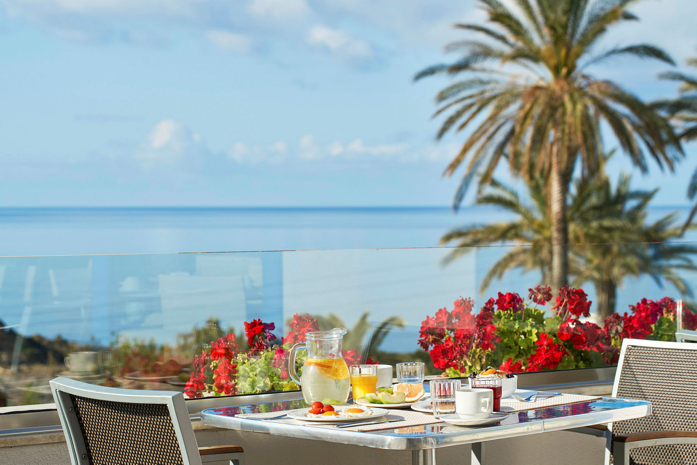 En morgenmadservering på en terrasse med havudsigt og røde blomster i fokus. (AI-genereret tekst)