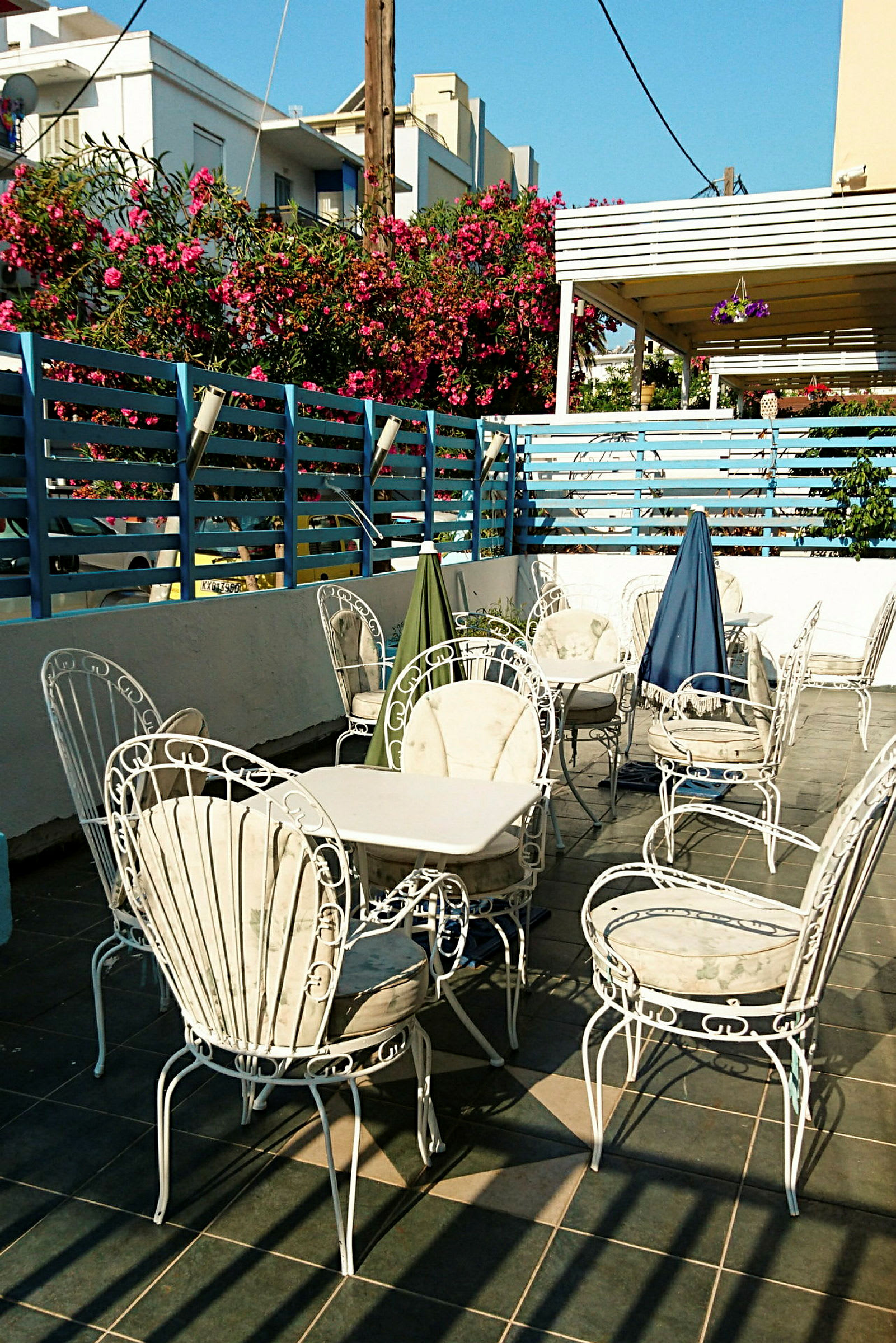 En terrasse med hvide havemøbler og borde, omgivet af et blåmalet hegn og blomstrende buske. (AI-genereret tekst)