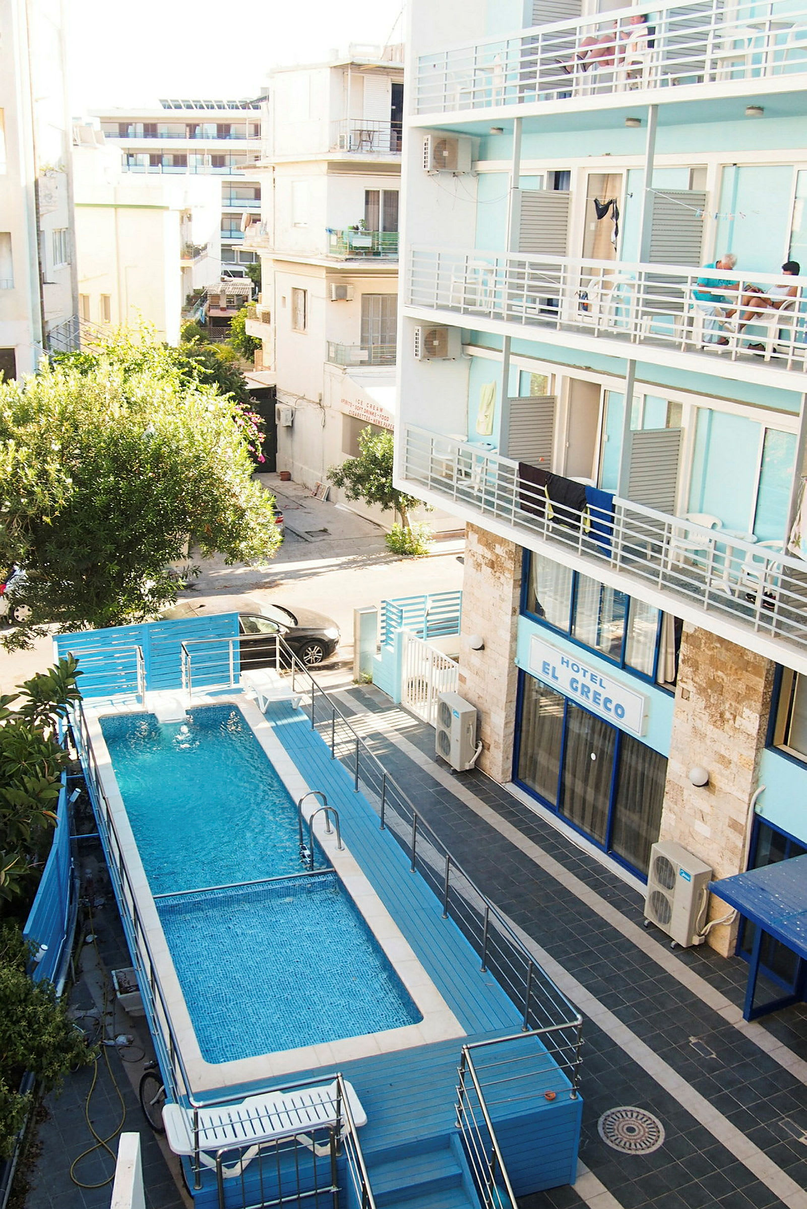 En swimmingpool med blåt vand omgivet af hegn, beliggende ved et hotel med blå facade. (AI-genereret tekst)