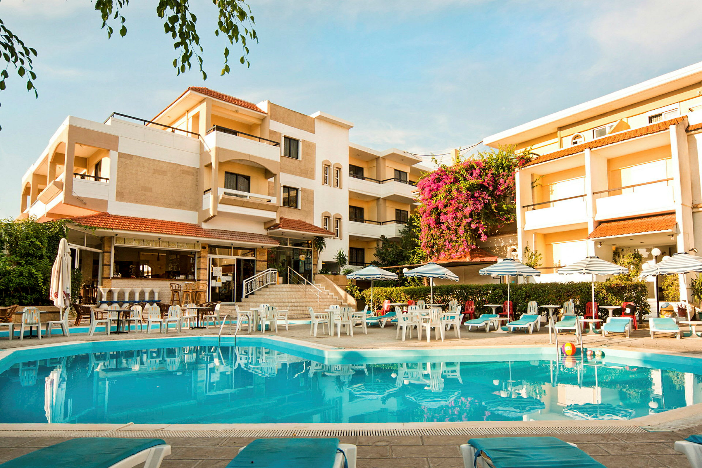 Hotelbygning med en pool i forgrunden, omgivet af liggestole og parasoller. (AI-genereret tekst)