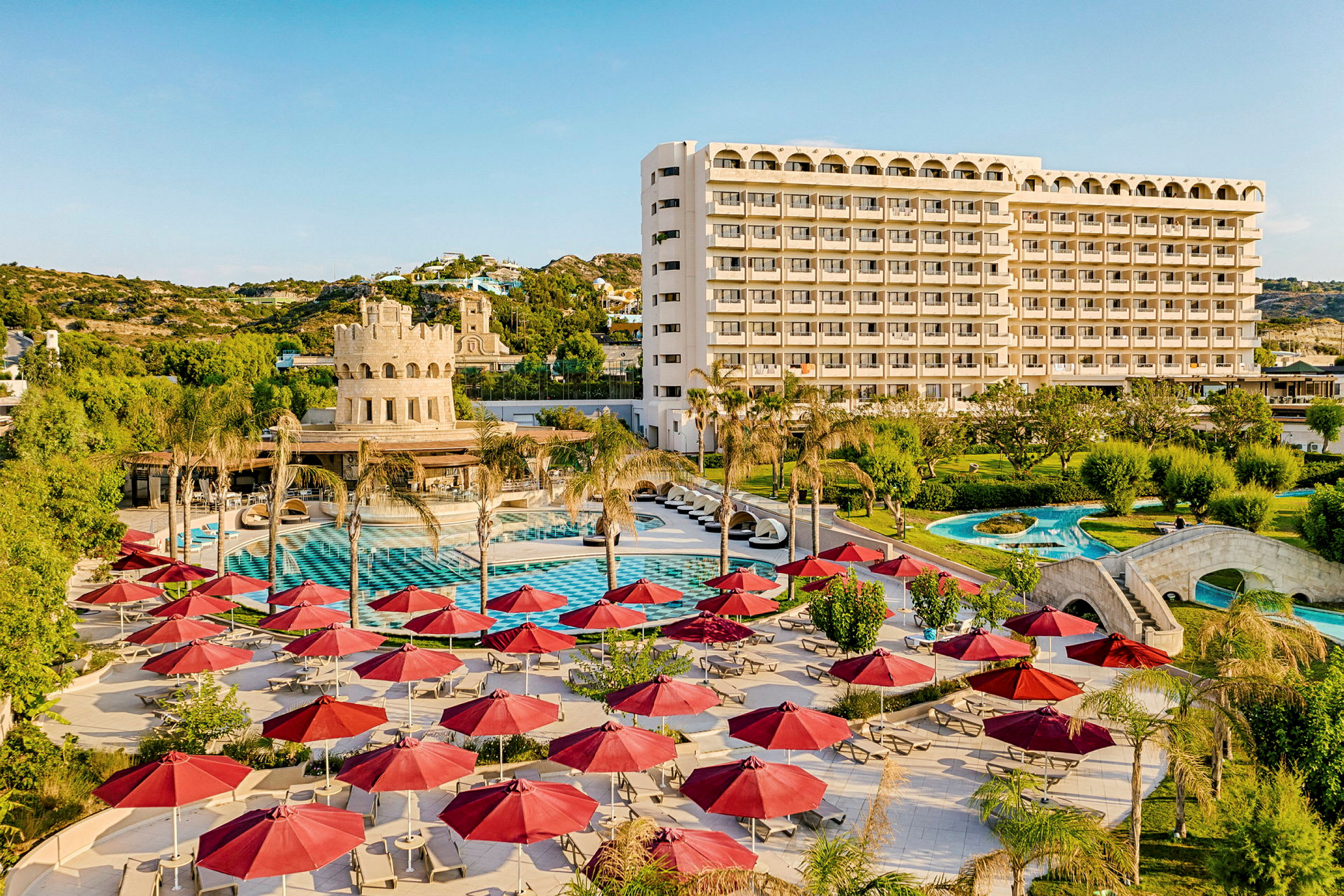 Hotelområde med pool, liggestole, røde parasoller og en større bygning i baggrunden. (AI-genereret tekst)