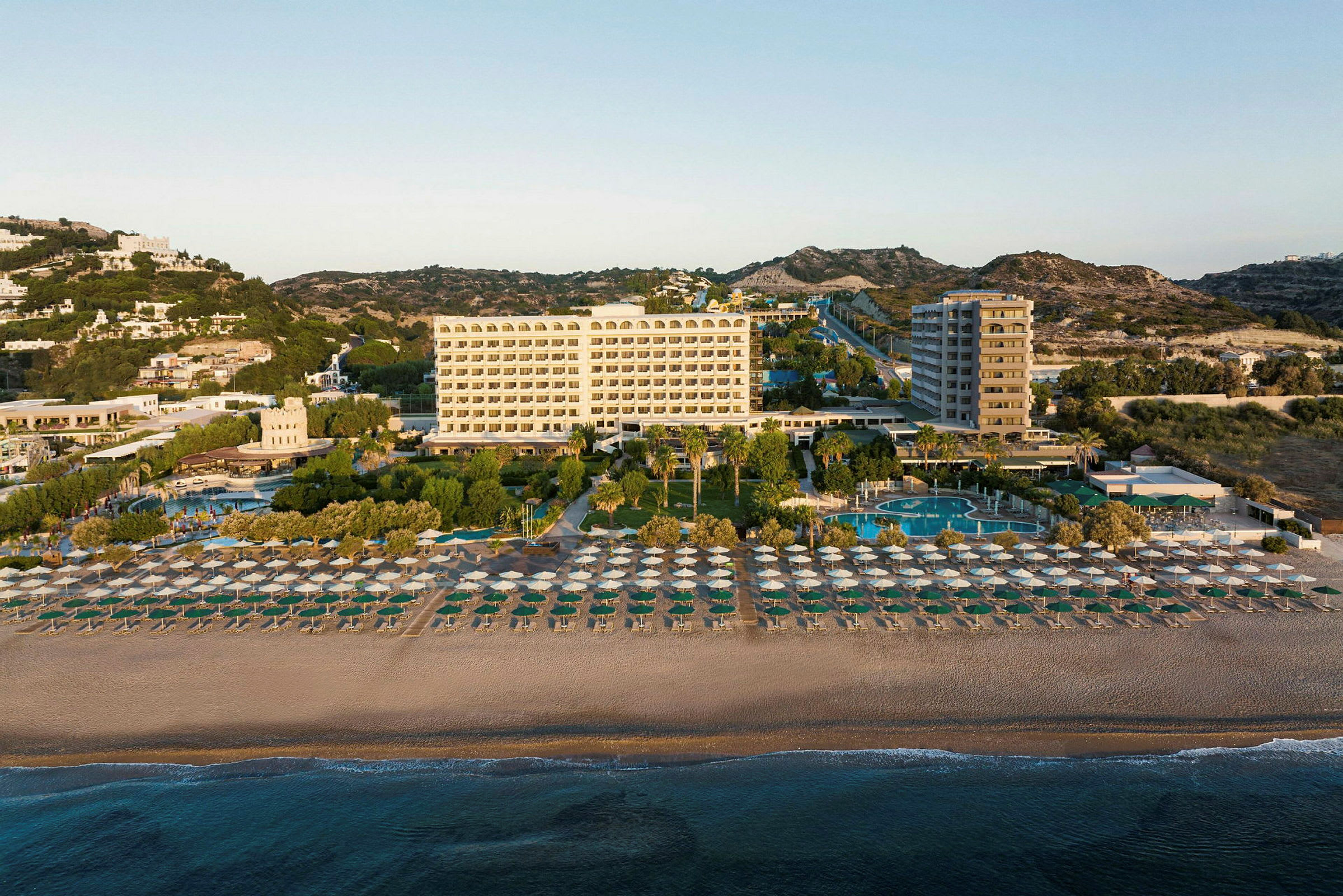 Et hotel med flere etager bag en sandstrand fyldt med parasoller, omgivet af træer og swimmingpools. (AI-genereret tekst)