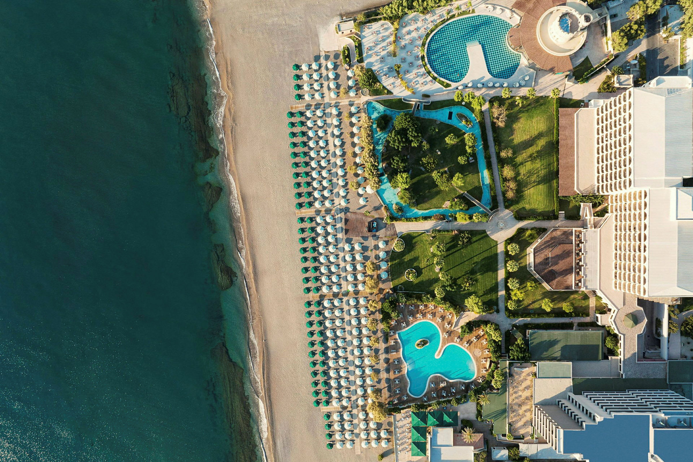 Luftfoto, der viser en strand med parasoller, poolområder og hotelbygninger. (AI-genereret tekst)