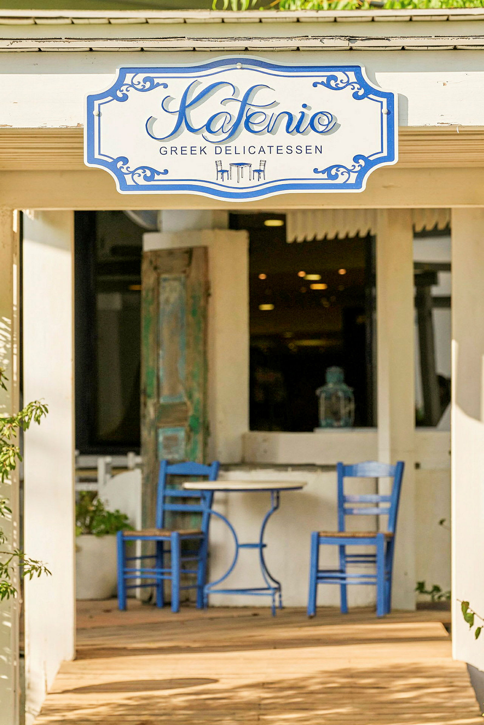To blå stole og et rundt bord står på en udendørsterrasse foran en bygning. En skilt over døren lyder "Kafenio Greek Delicatessen". (AI-genereret tekst)