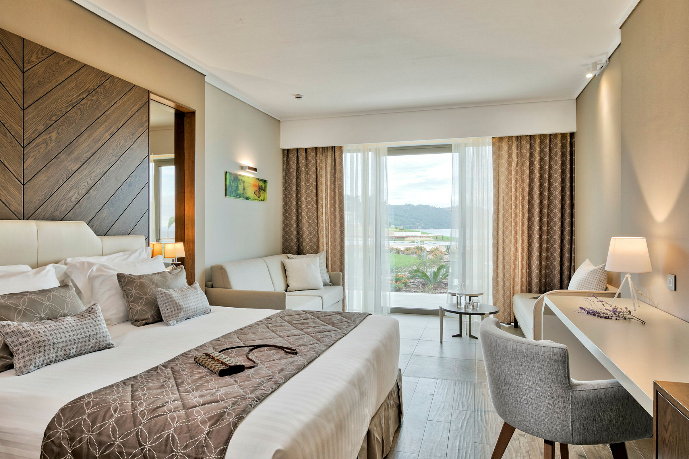 Et moderne hotelværelse med en stor seng, en sofa og et skrivebord med en lampe ved siden af. Vinduesudsigten viser et landskab. (AI-genereret tekst)