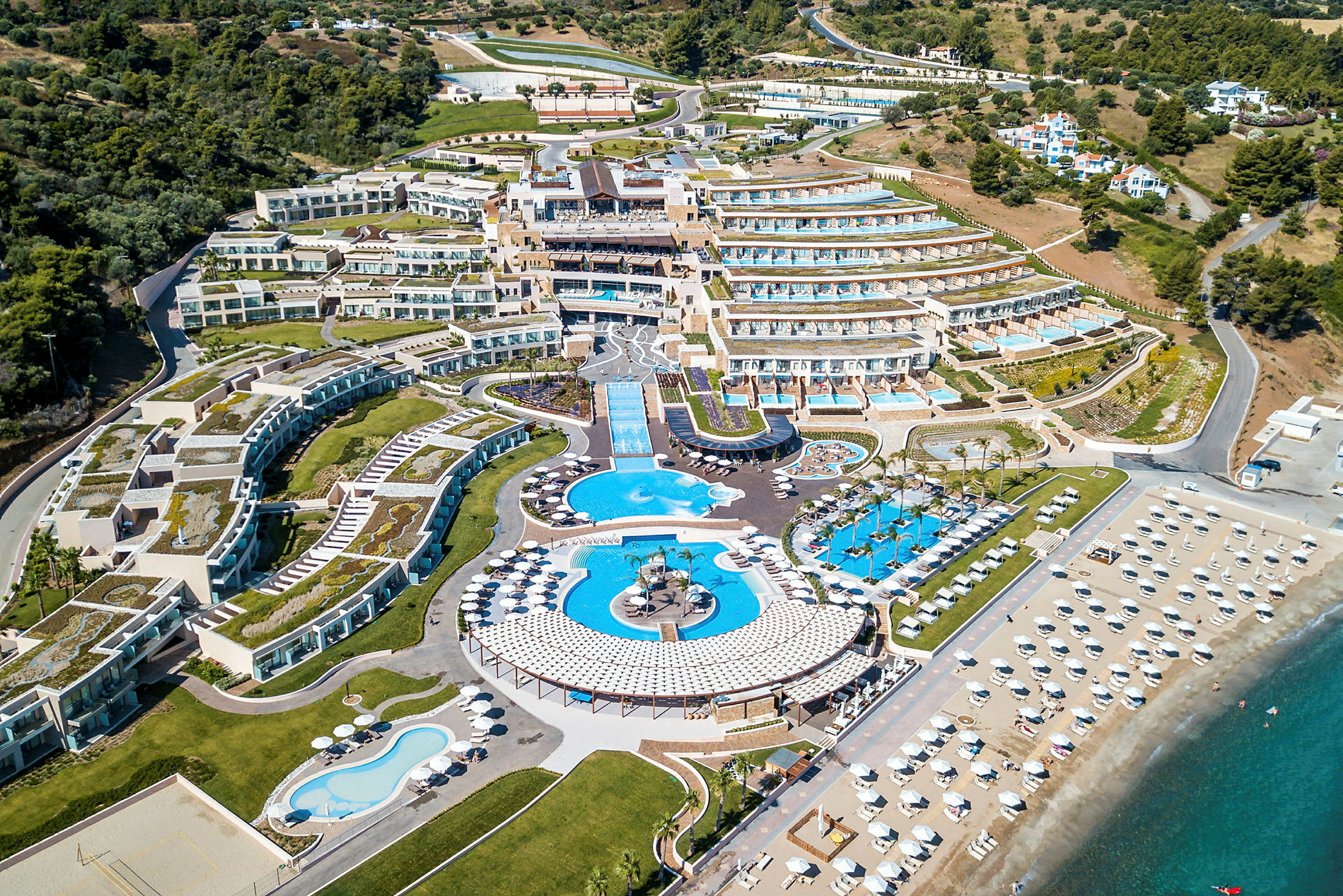 Luftfoto af et stort feriested med terrasserede bygninger, flere swimmingpools og en bred sandstrand med parasoller. (AI-genereret tekst)