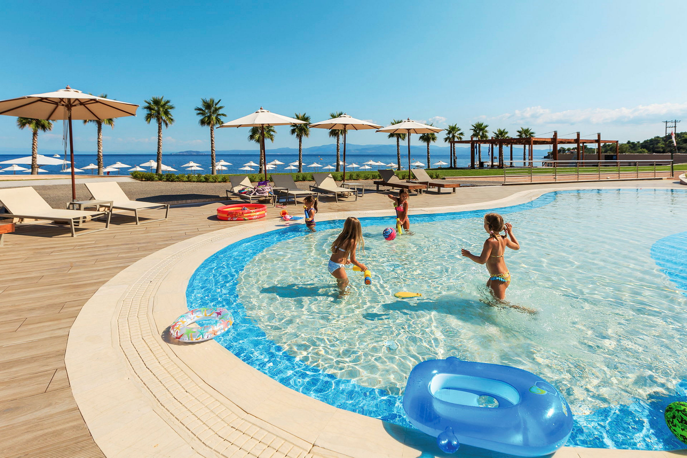 Børn leger i en lav pool ved en resort nær havet. Liggestole og parasoller er placeret rundt om poolområdet. (AI-genereret tekst)