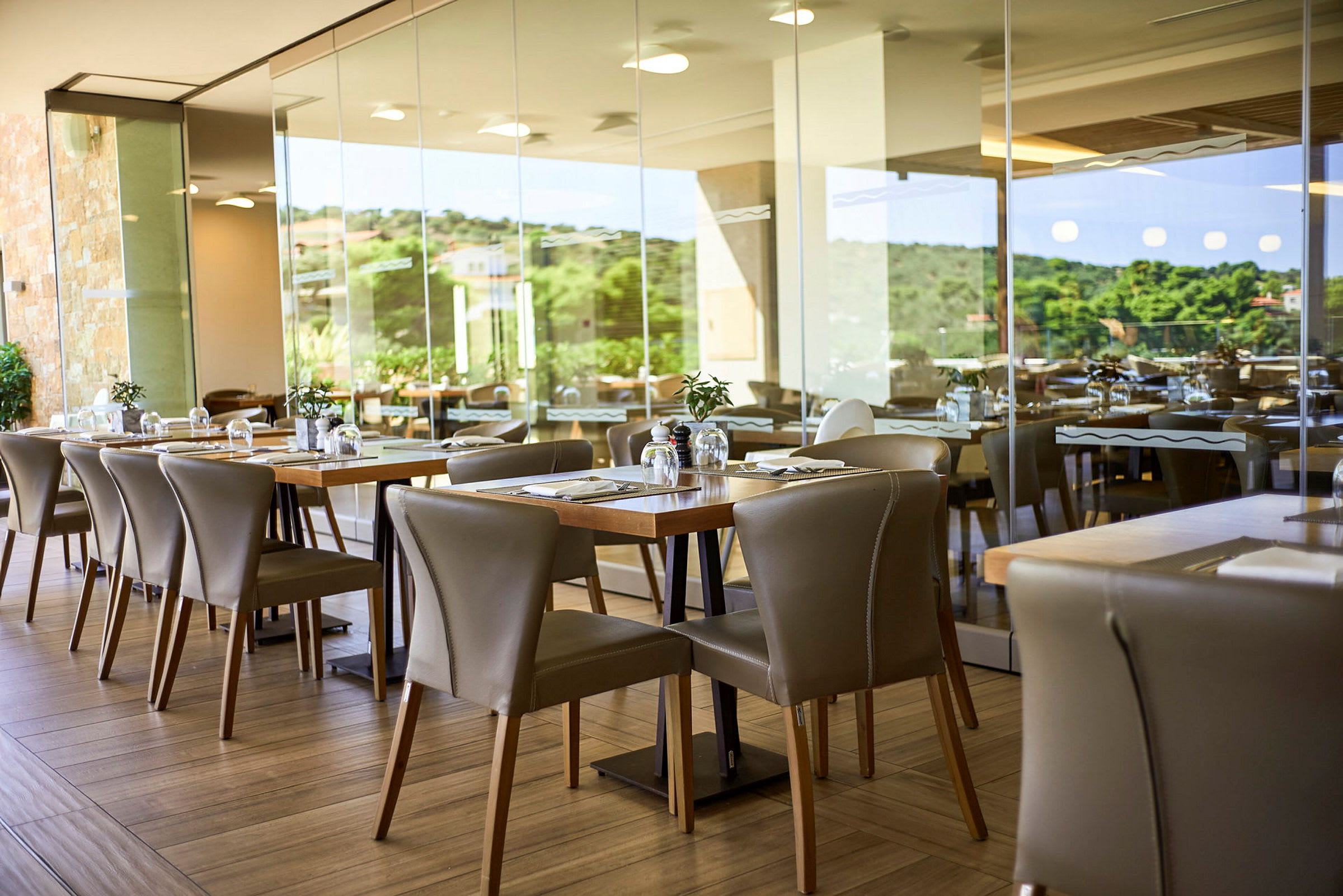 Et moderne restaurantområde med borde og stole samt udsigt gennem glasvægge mod grønne områder. (AI-genereret tekst)