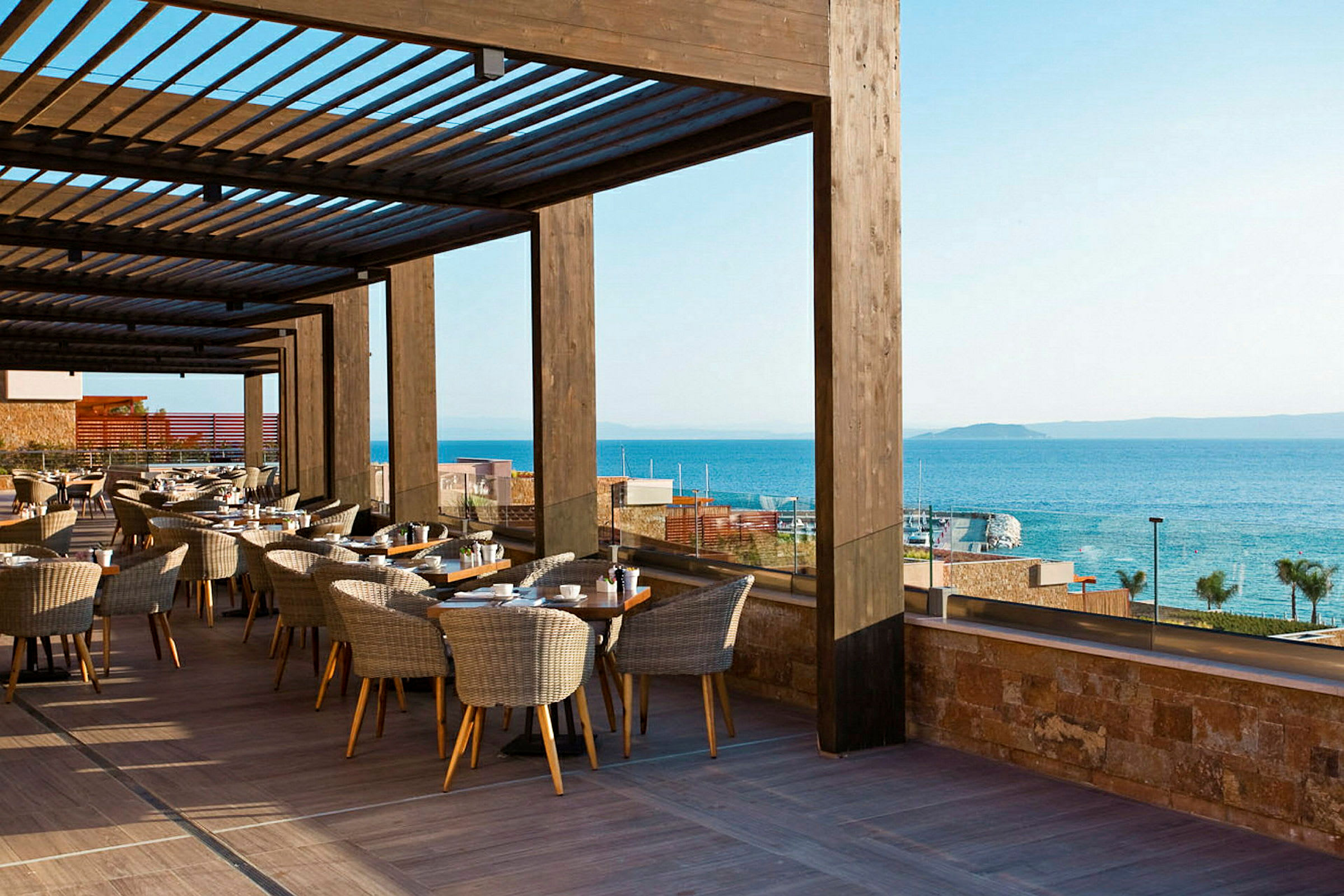 En terrasse med borde og stole under en træpergola med udsigt over havet i baggrunden. (AI-genereret tekst)
