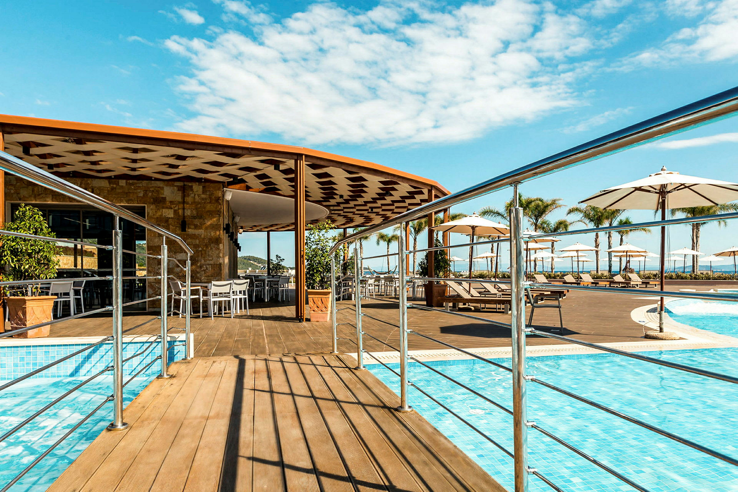 En udendørs pool med en træterrasse, parasoller og solsenge. I baggrunden ses en bygning med udendørs serveringsområde. (AI-genereret tekst)