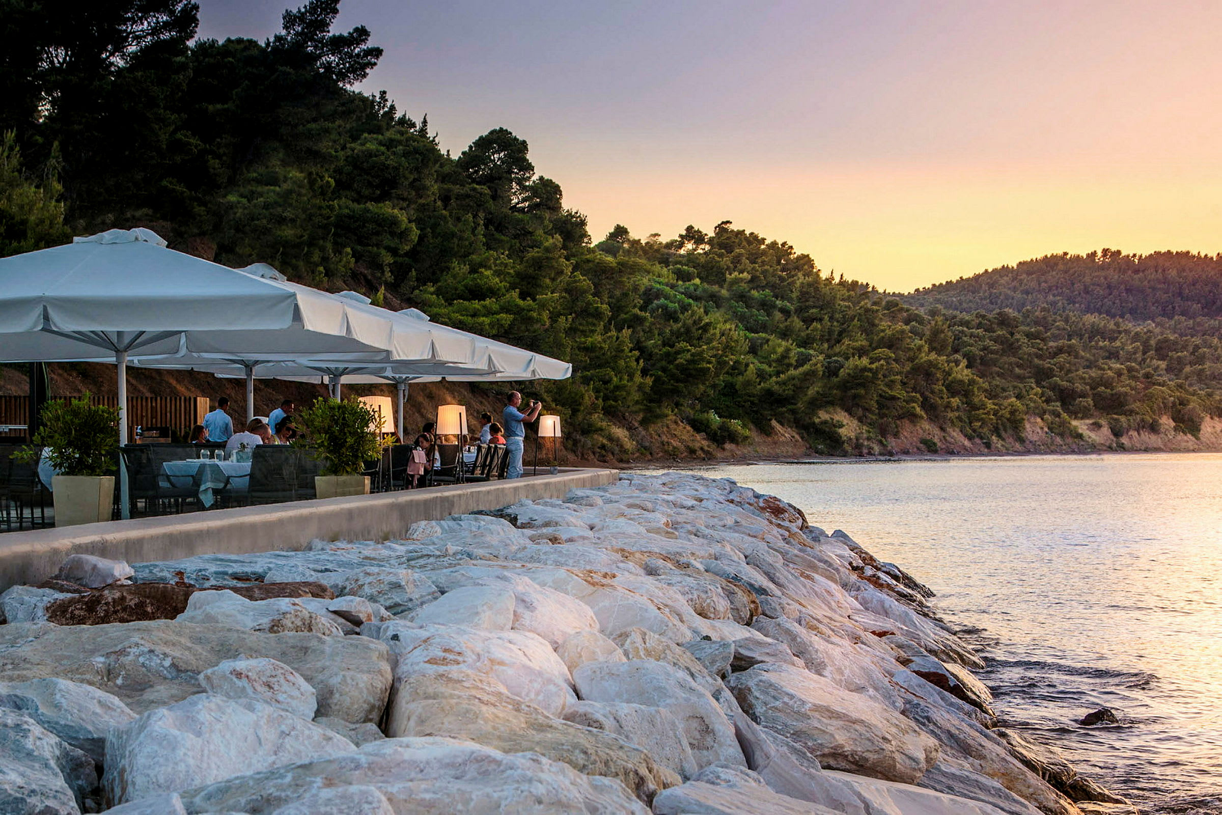 En restaurant med hvide parasoller ved stranden, omgivet af grøntområde og vand med solnedgang. (AI-genereret tekst)