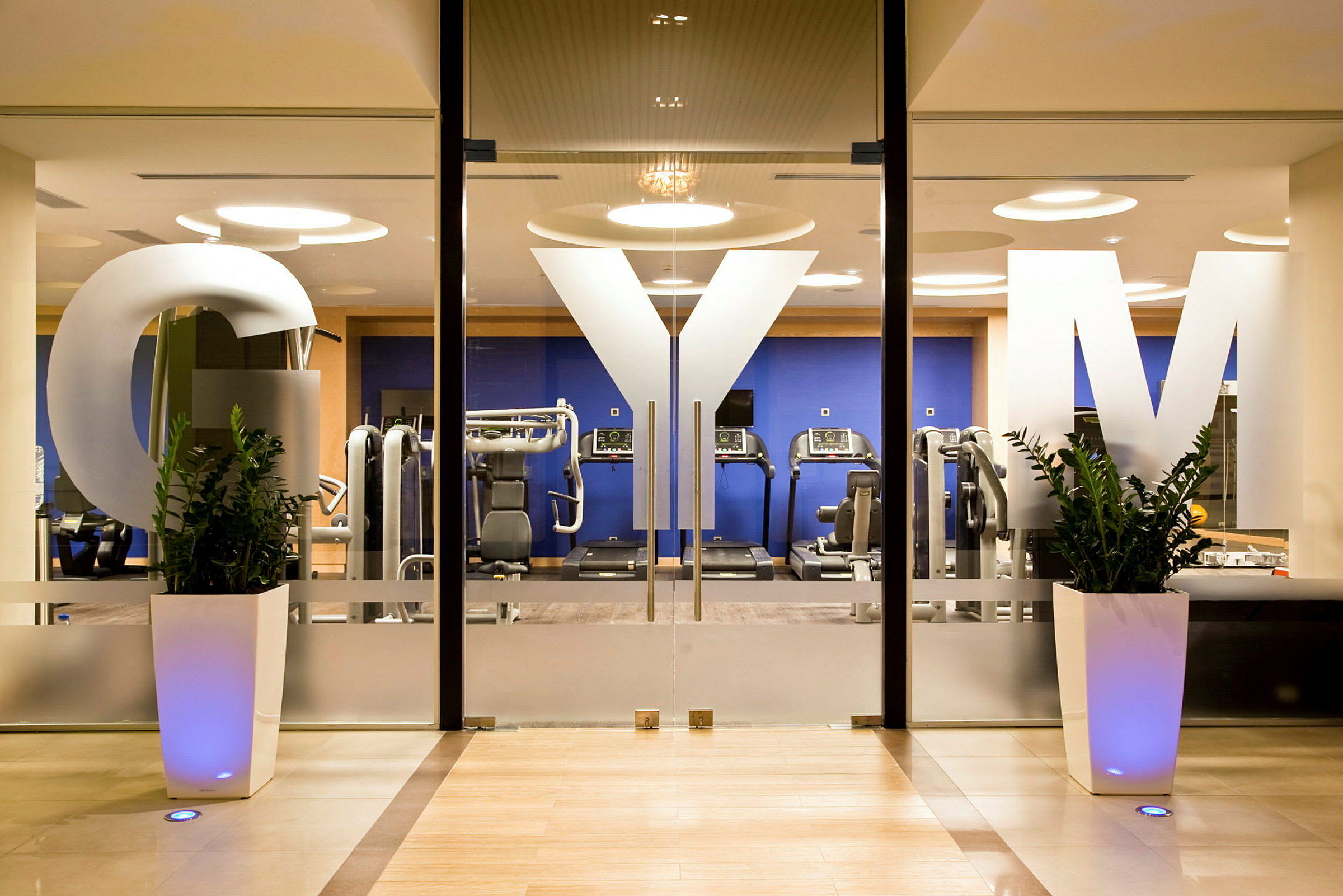 En indgang til et fitnesscenter med glasdøre dekoreret med bogstaverne "GYM" samt synlige maskiner og træningsudstyr i baggrunden. (AI-genereret tekst)