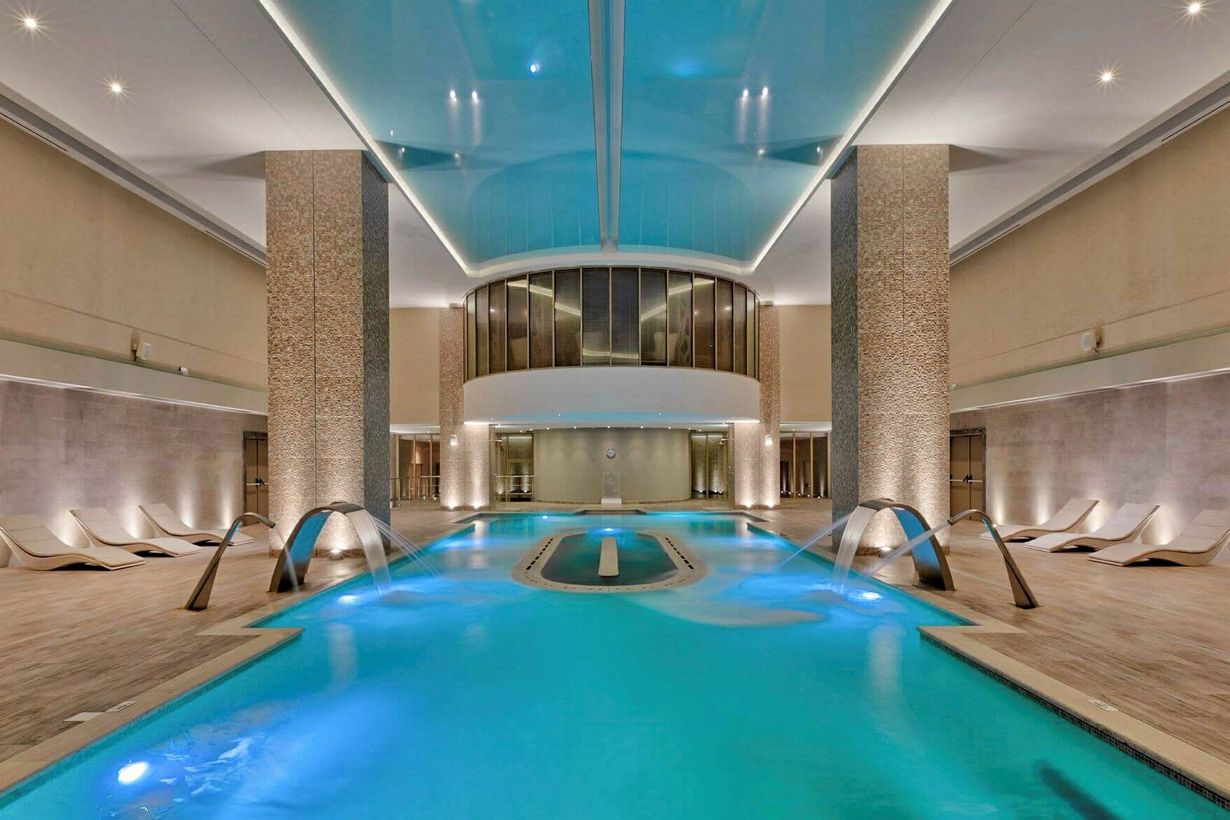 Et moderne indendørs spa med en oplyst pool, vandstråler og komfortable liggestole på siderne. (AI-genereret tekst)