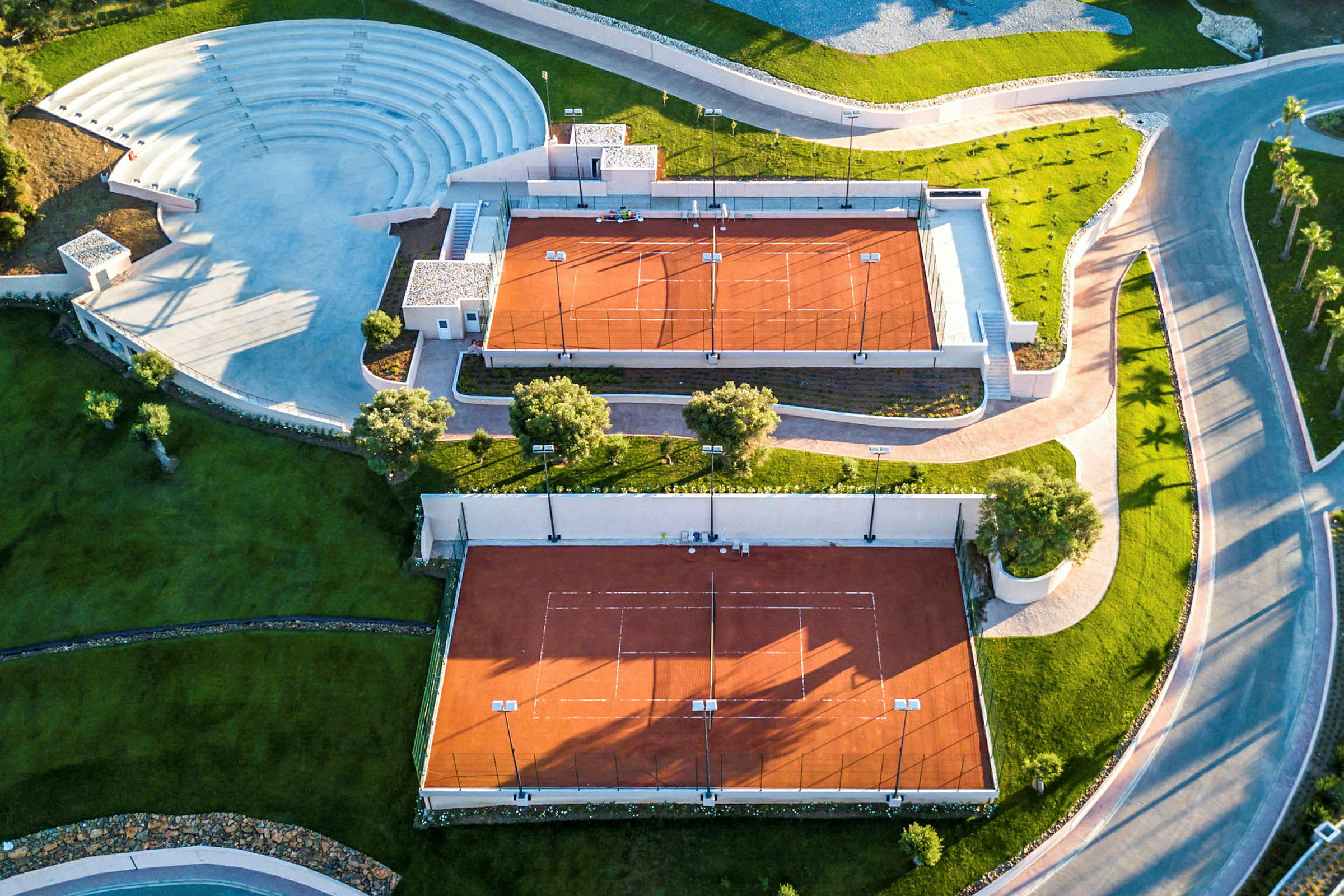 Luftfoto af to tennisbaner med rød belægning og et tilstødende udendørs amfiteater. (AI-genereret tekst)