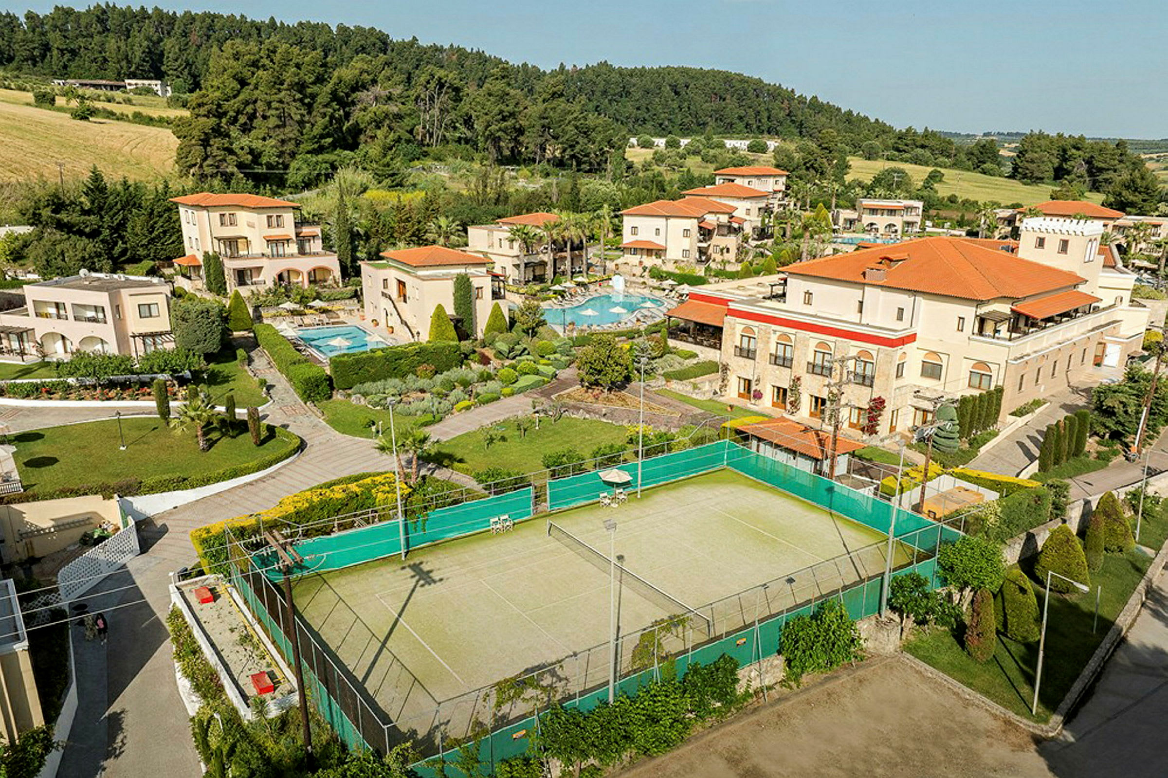 Luftfoto af et feriekompleks med swimmingpools, haver og en tennisbane, omgivet af grønne områder og skov. (AI-genereret tekst)