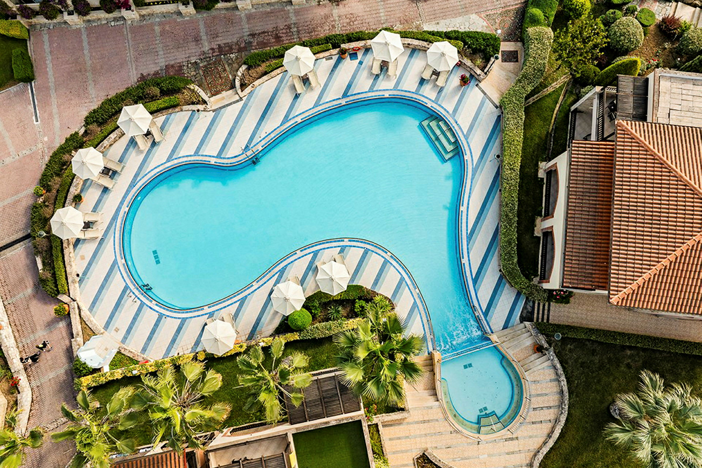 Et vue fra oven af en swimmingpool omgivet af parasoller og grønne områder tæt på en bygning med tag. (AI-genereret tekst)