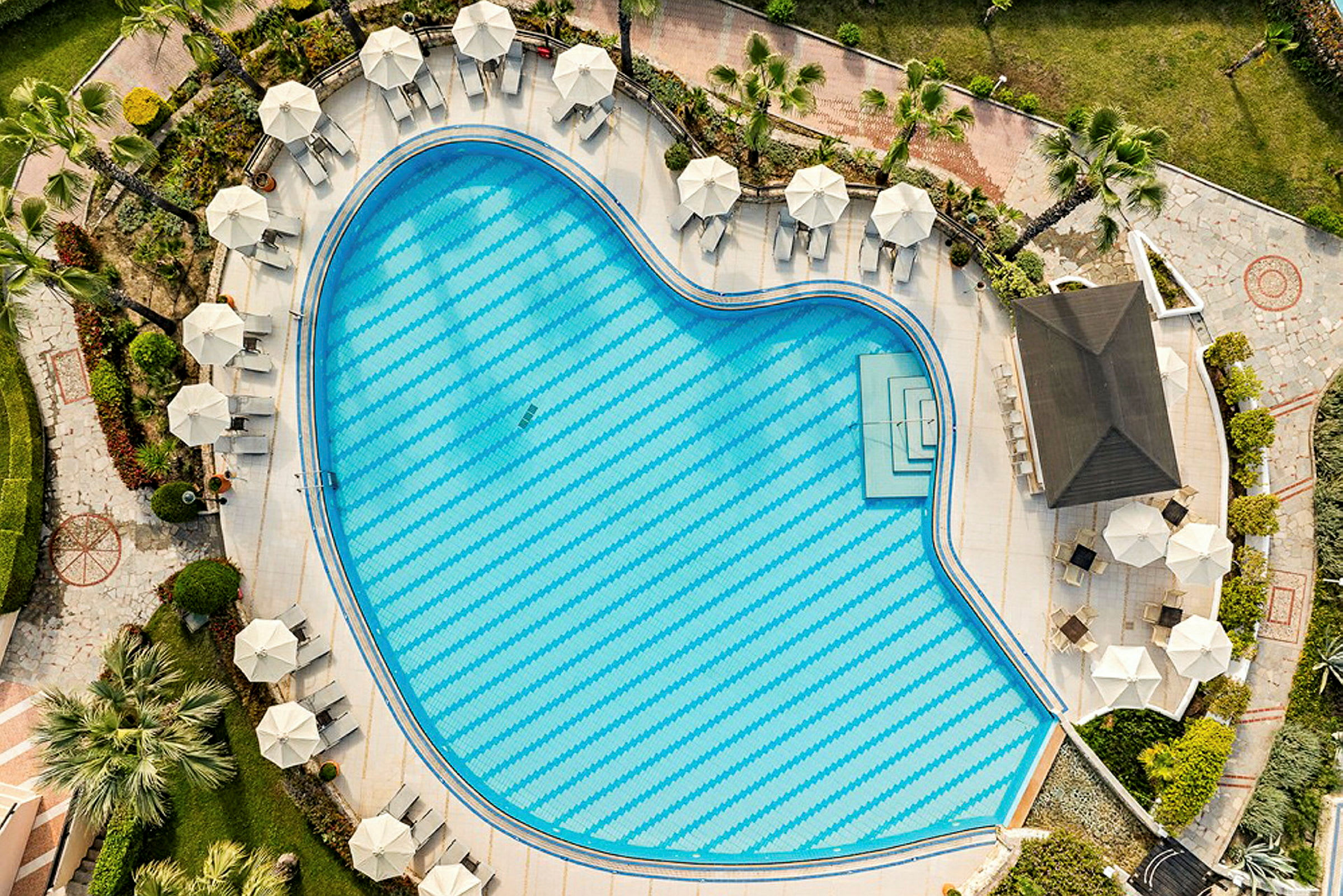 Luftfoto af en hjerteformet pool omgivet af parasoller og liggestole placeret i et velholdt poolområde. (AI-genereret tekst)